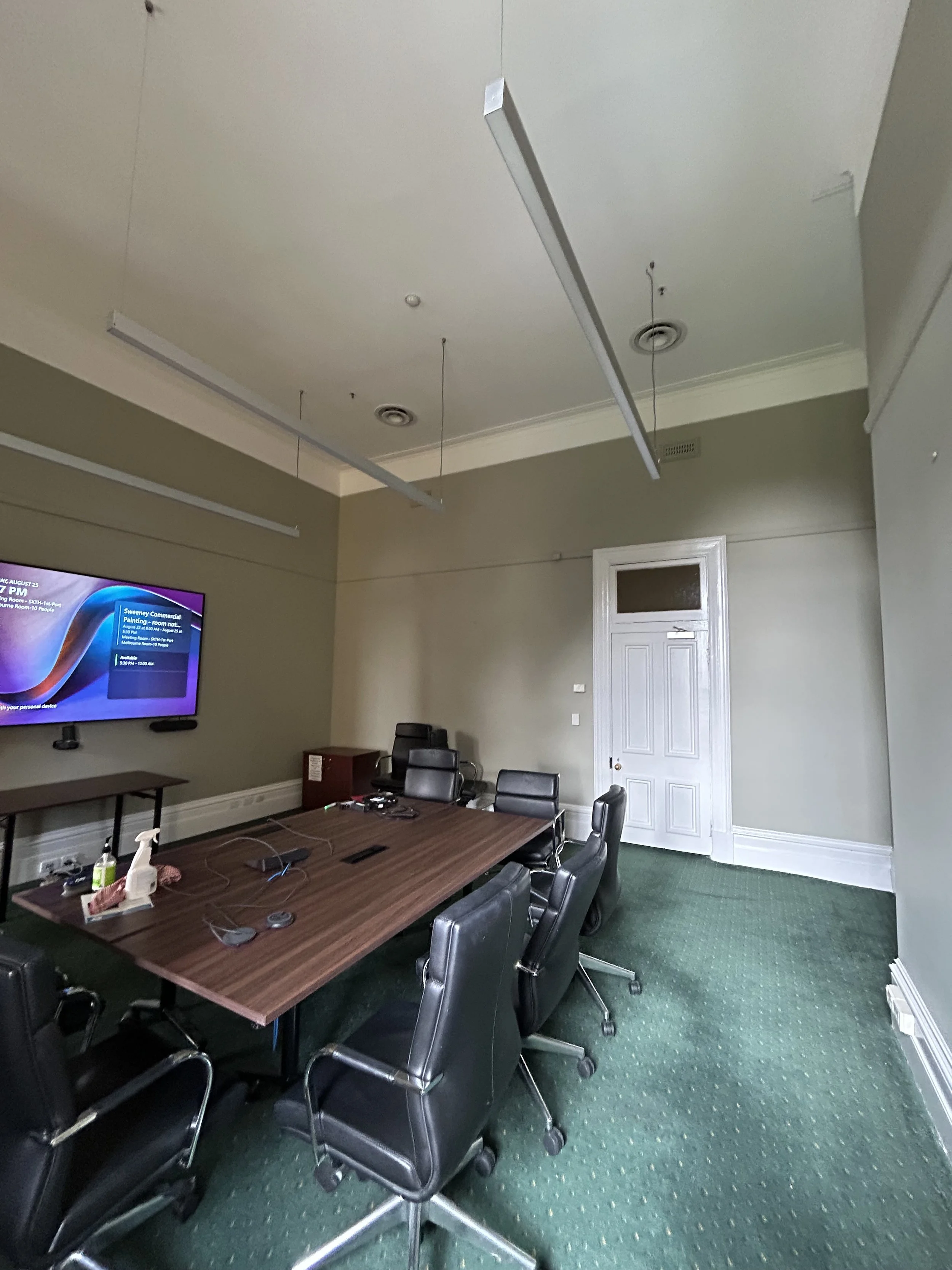 Empty conference room with a large wooden table, black office chairs, a wall-mounted screen displaying a presentation, and ceiling-mounted light fixtures.