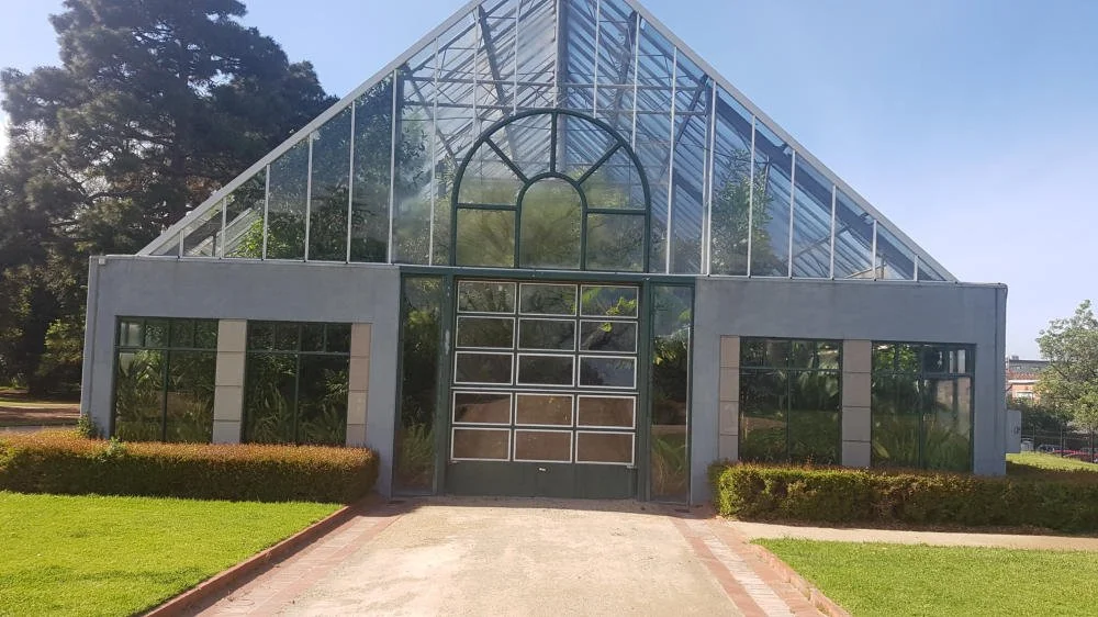 A large greenhouse with glass walls and a peaked roof, surrounded by green grass and shrubbery, with trees and a clear sky in the background.