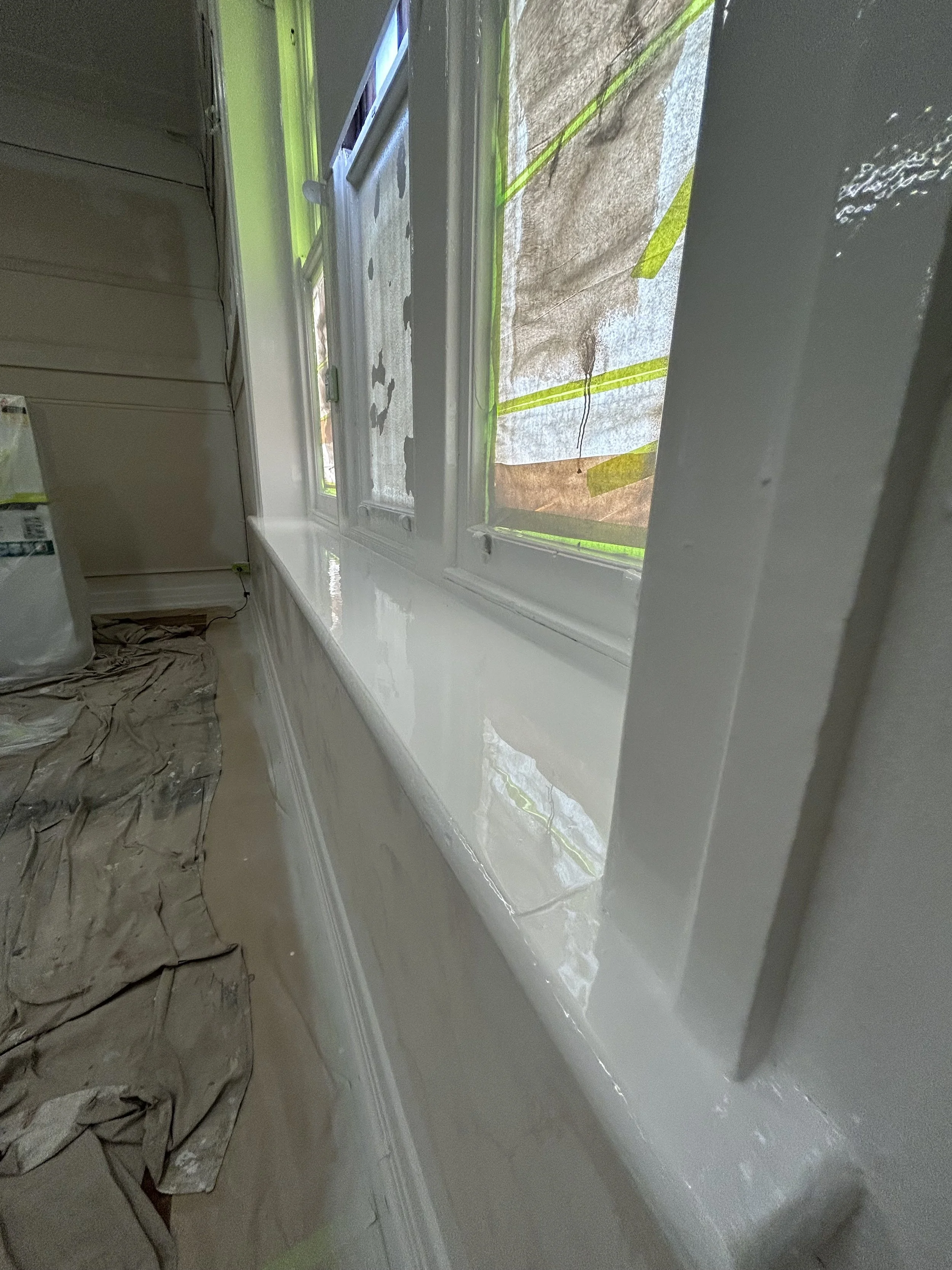 Close-up of a freshly painted white window sill next to a window with partially covered glass, and a beige sheet or tarp on the floor beneath the window.