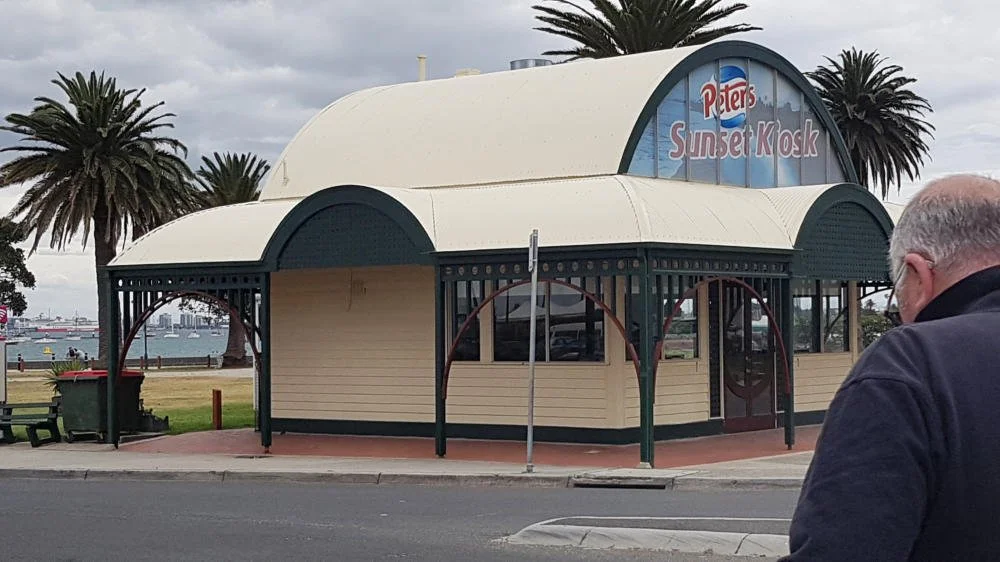 A small kiosk named Peters Sunset Kiosk with a curved roof, located near a waterfront with palm trees in the background.
