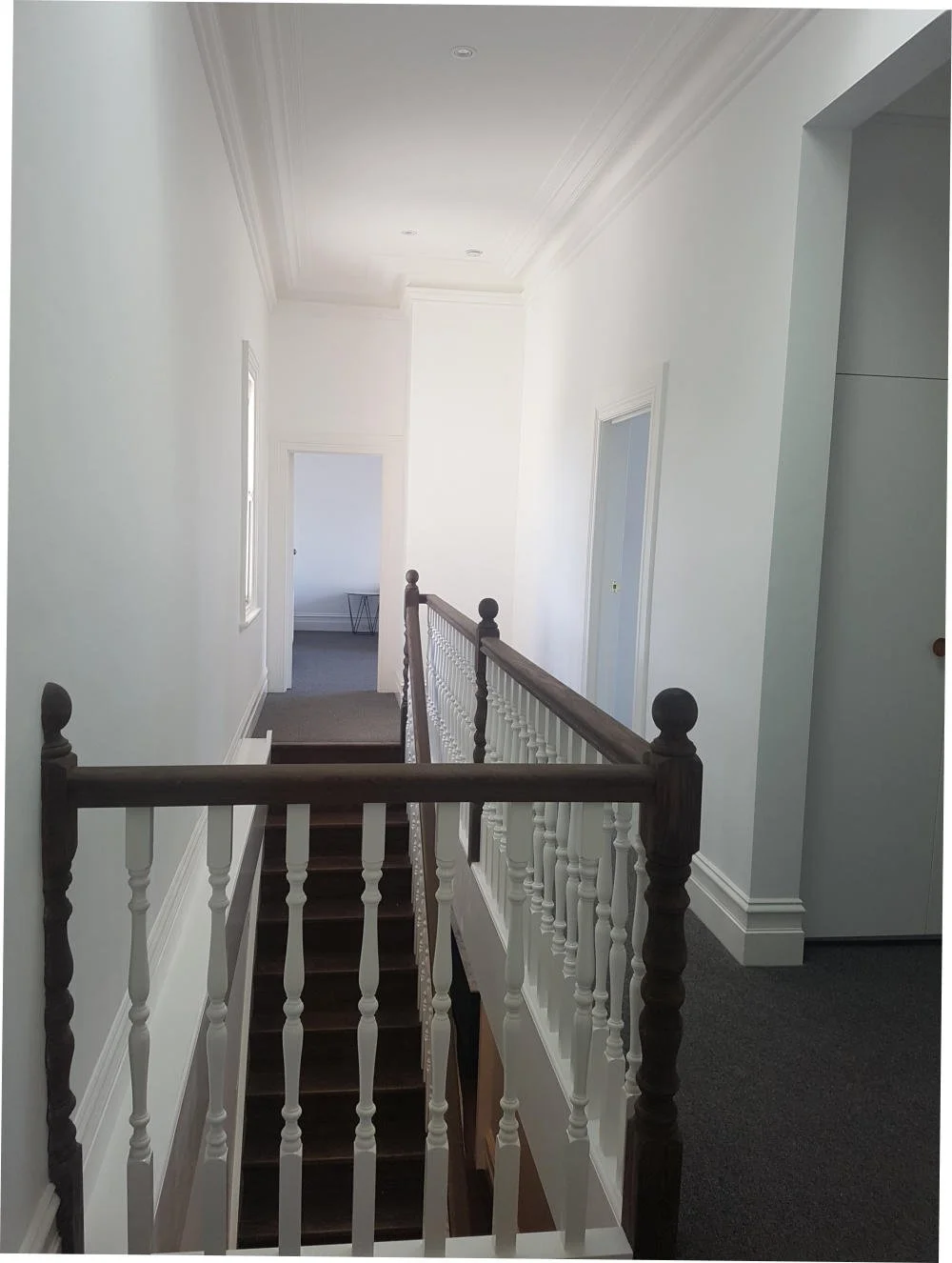 Interior of a bright, white hallway with a staircase and wooden handrail, leading to a room at the end.