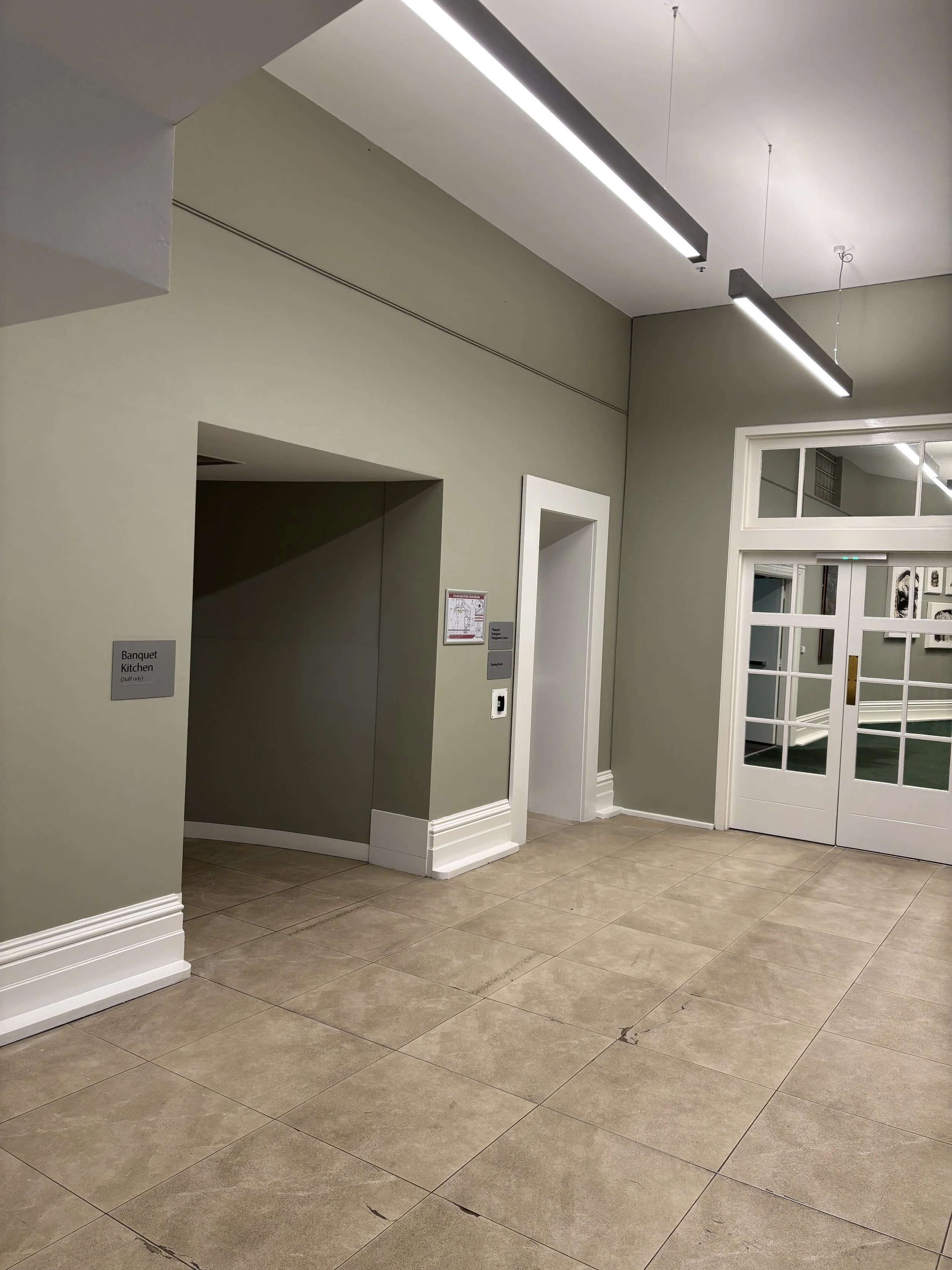 A hallway with beige floor tiles, green walls, and white trim. There are glass double doors on the right and a sign indicating a banquet kitchen on the left. Fluorescent lights hang from the ceiling.