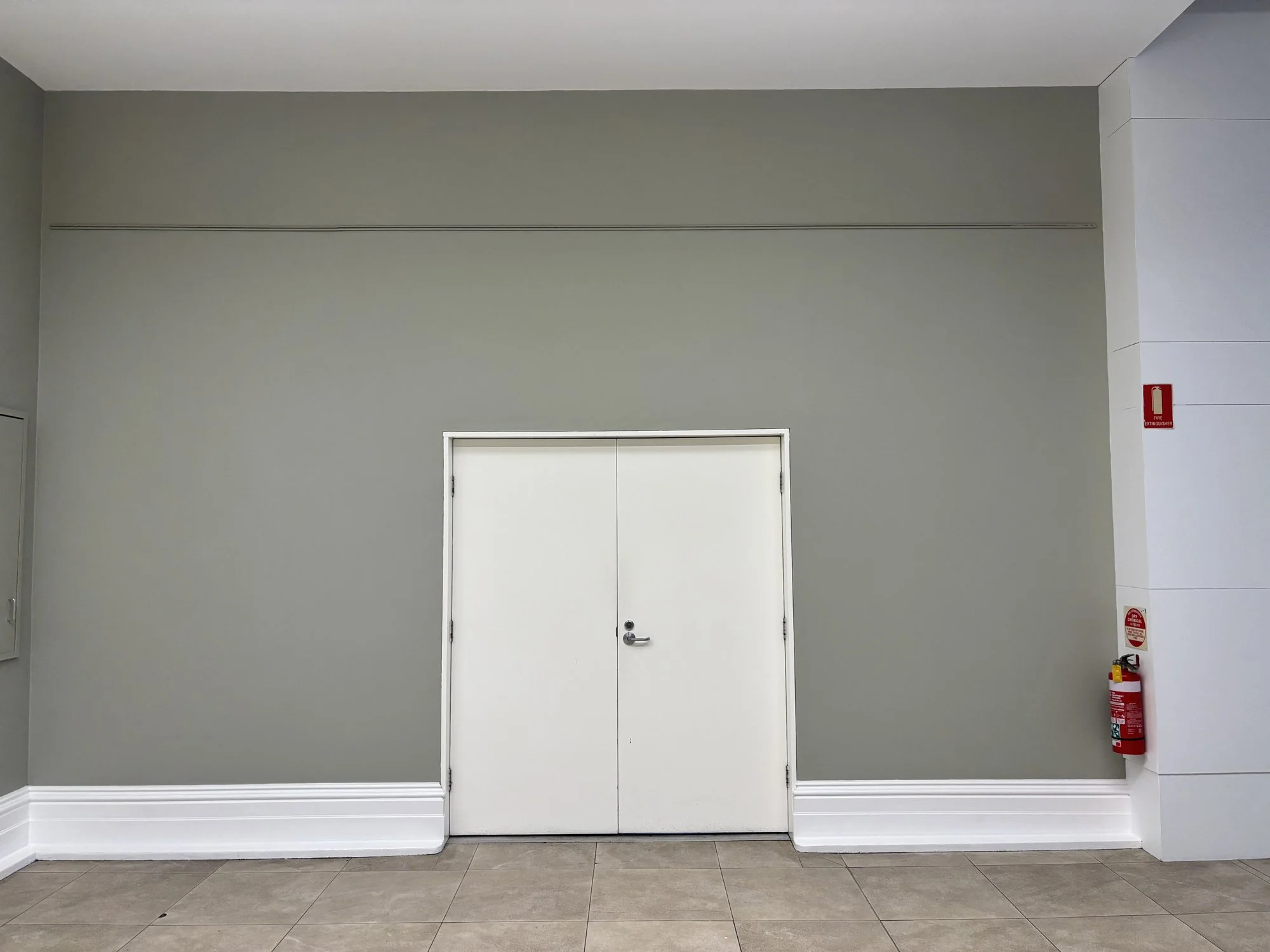 A plain gray wall with a white double door in the center and white baseboards at the bottom. To the right, there is a red fire extinguisher mounted on the wall.