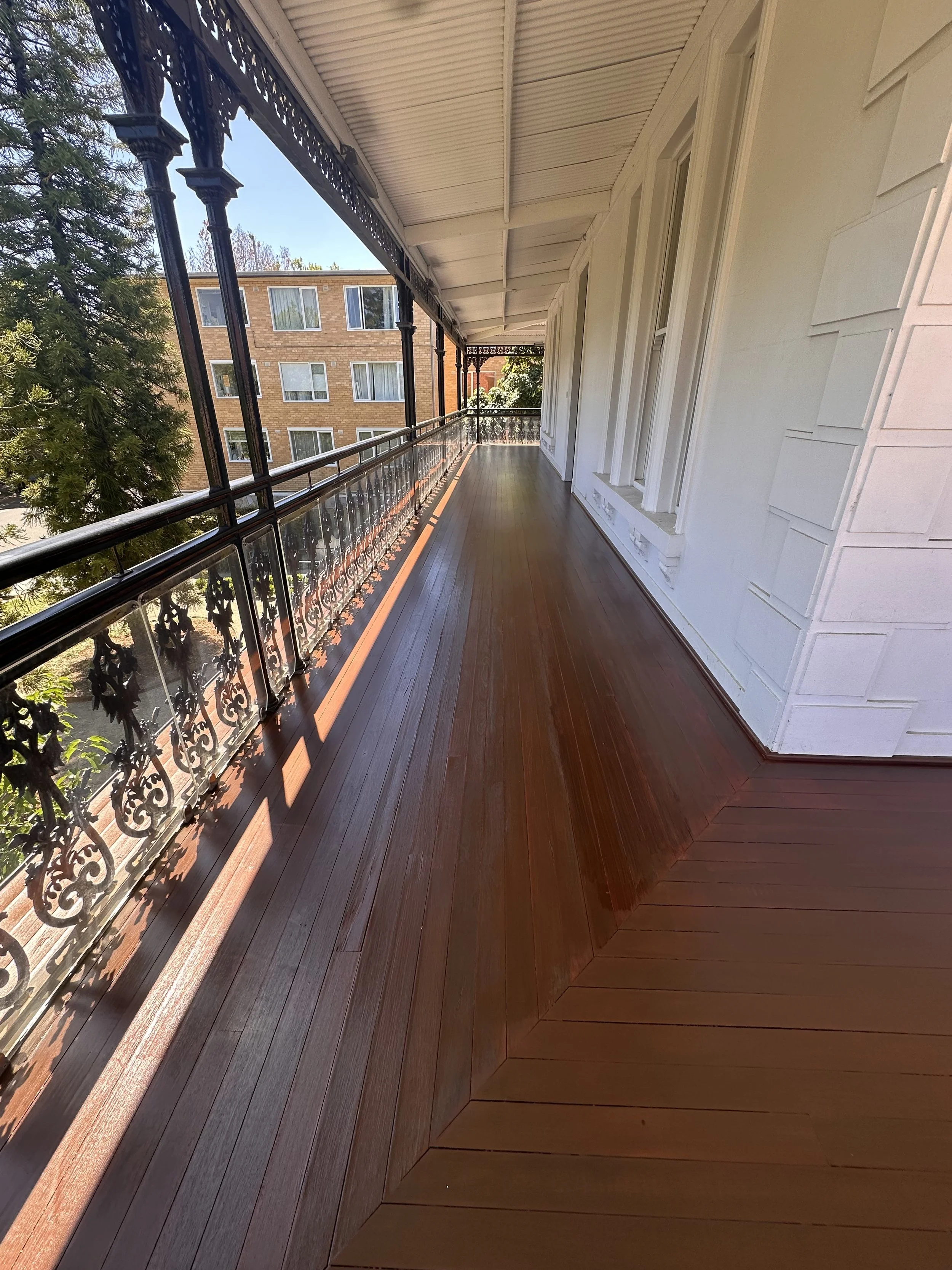 Empty porch with polished wooden floor, decorative black iron railing, white exterior wall, and windows, in front of residential building surrounded by trees.