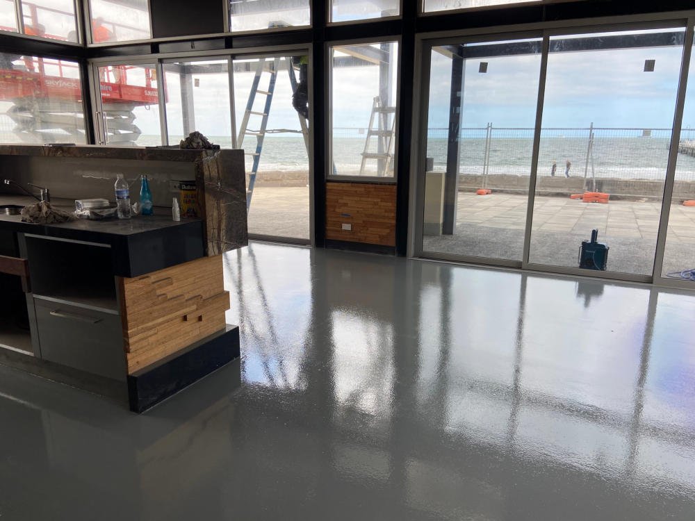 Interior of a building under construction with a view of the beach and ocean outside through large glass windows and doors.