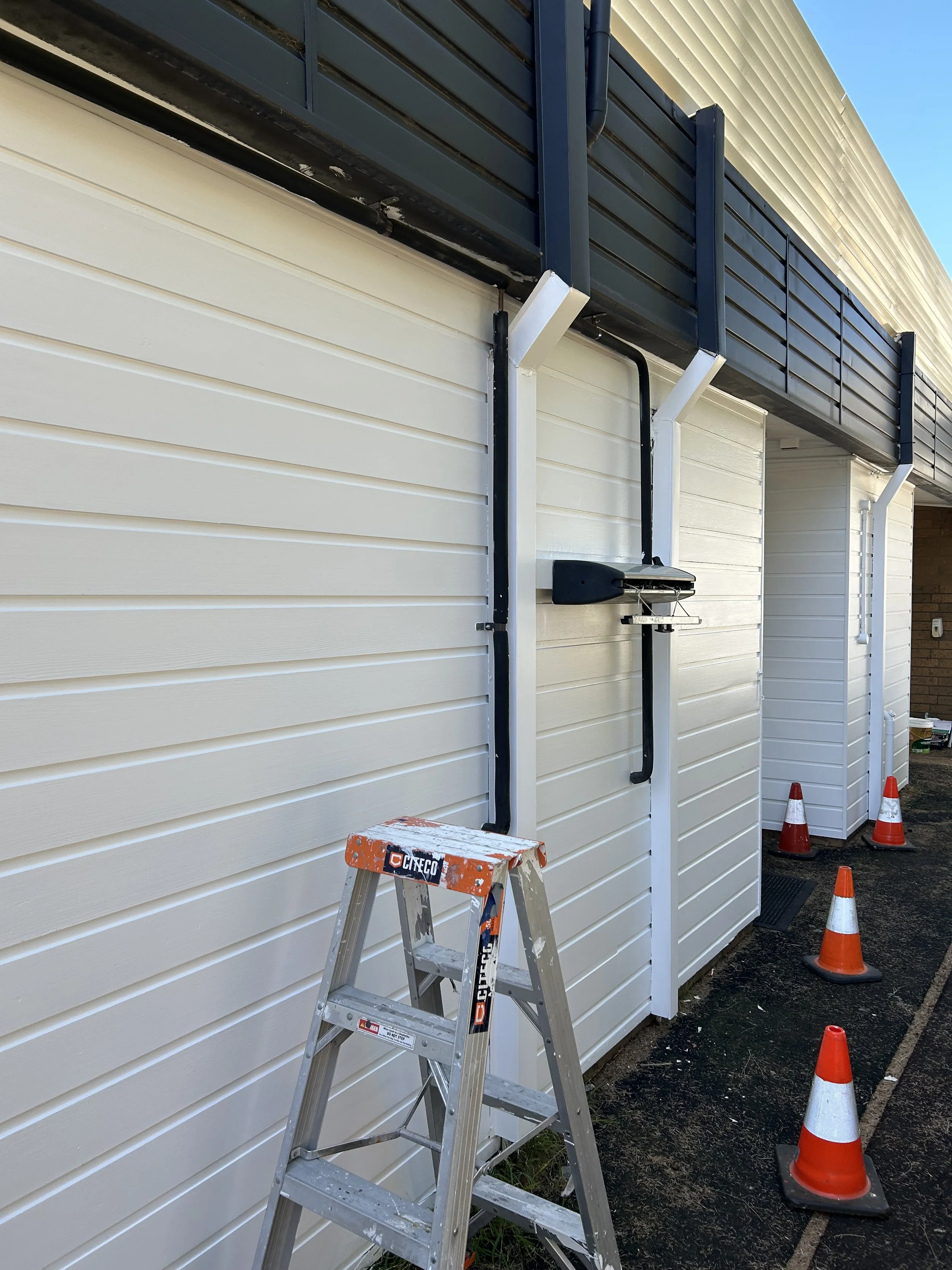 Construction site with a stepladder and orange traffic cones outside a building with white and dark siding, electrical conduit, and a black motorized valve or switch mounted on the wall.