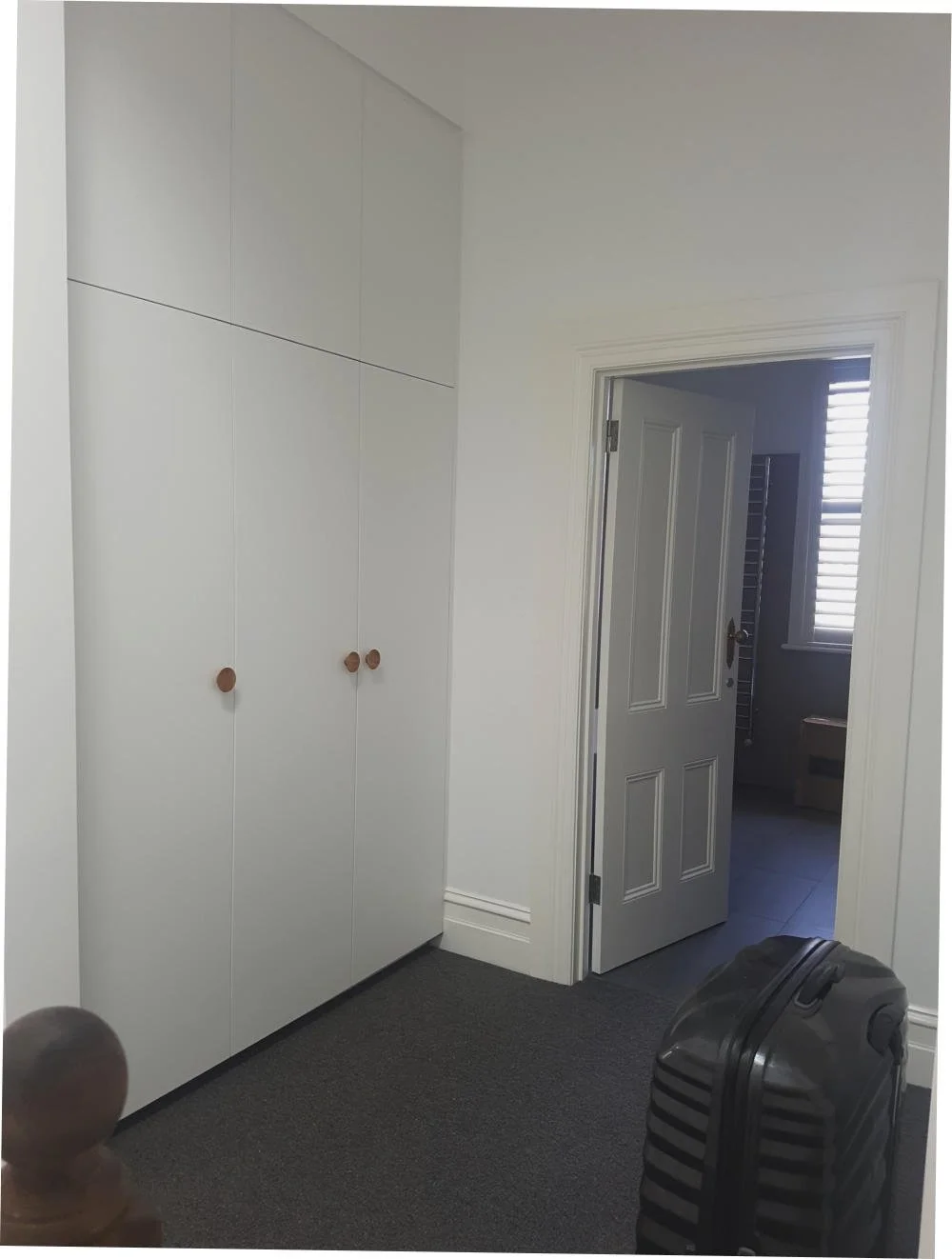 A white built-in closet with three doors and round wooden knobs, an open door leading to a room with a window and blinds, a black suitcase on dark carpet, and part of a wooden bedpost in the foreground.