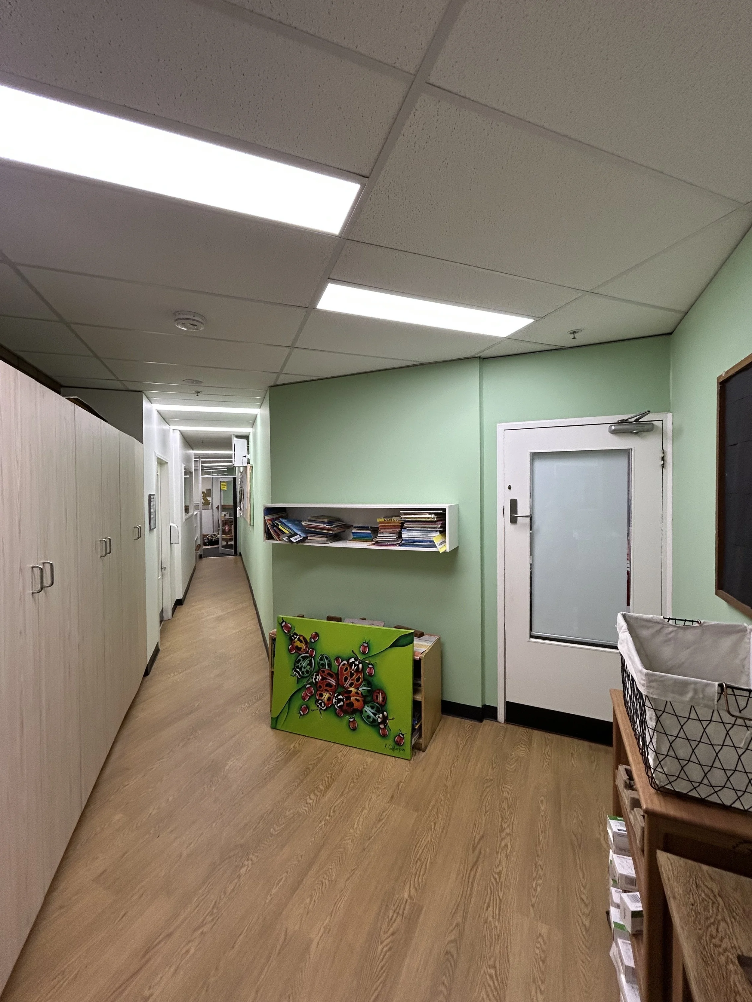 A hallway in an office or classroom with lockers on the left, a light green wall with a shelf full of books and magazines, and a door with a frosted glass window. 