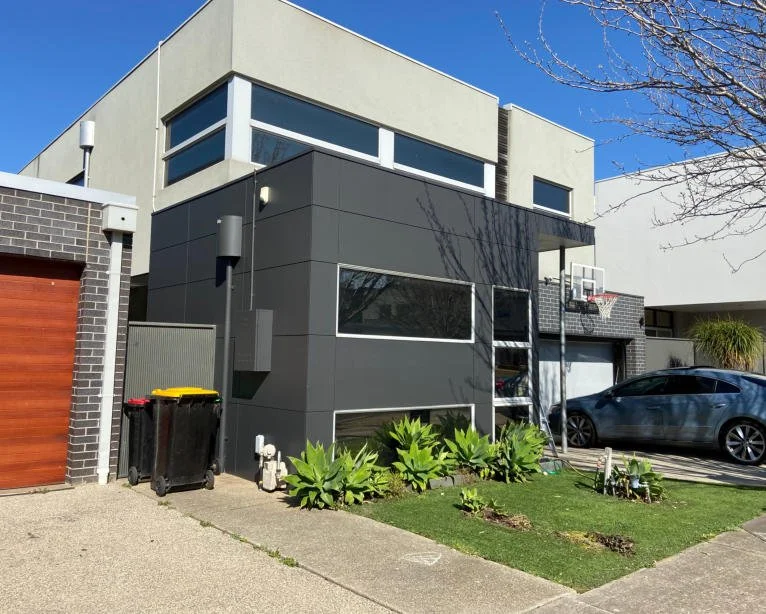 Modern two-story house with gray and white exterior, large windows, a garage, and a parked gray car in the driveway. A basketball hoop and small garden with green plants are visible in the front yard.
