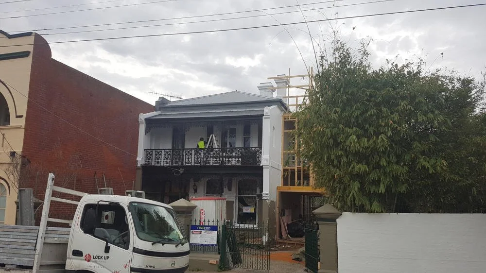 Two-story building under renovation with workers on the balcony, a truck parked in front, and a large tree next to a white fence.