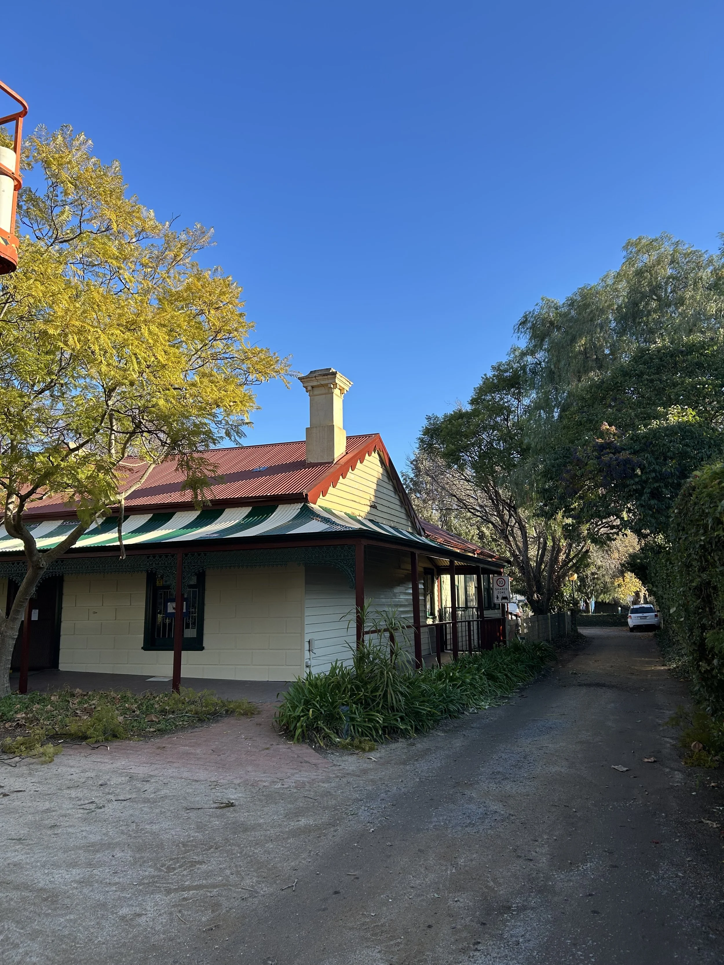 A house with a red roof, light-colored walls, and a chimney, surrounded by trees and a dirt road, under a clear blue sky.