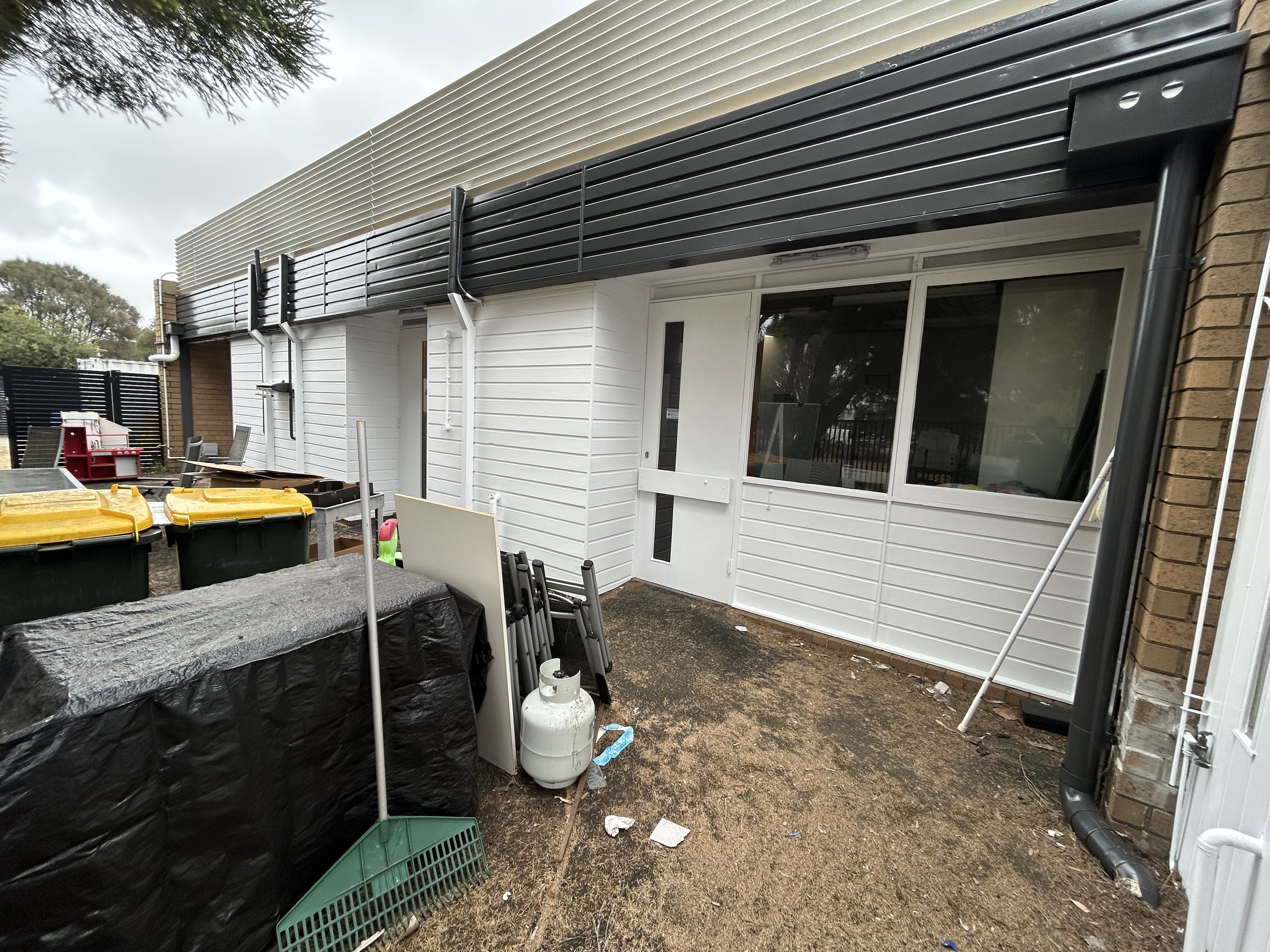Exterior view of a building with white and black siding, with a window and door, and clutter including garbage bins, a propane tank, and various outdoor furniture.