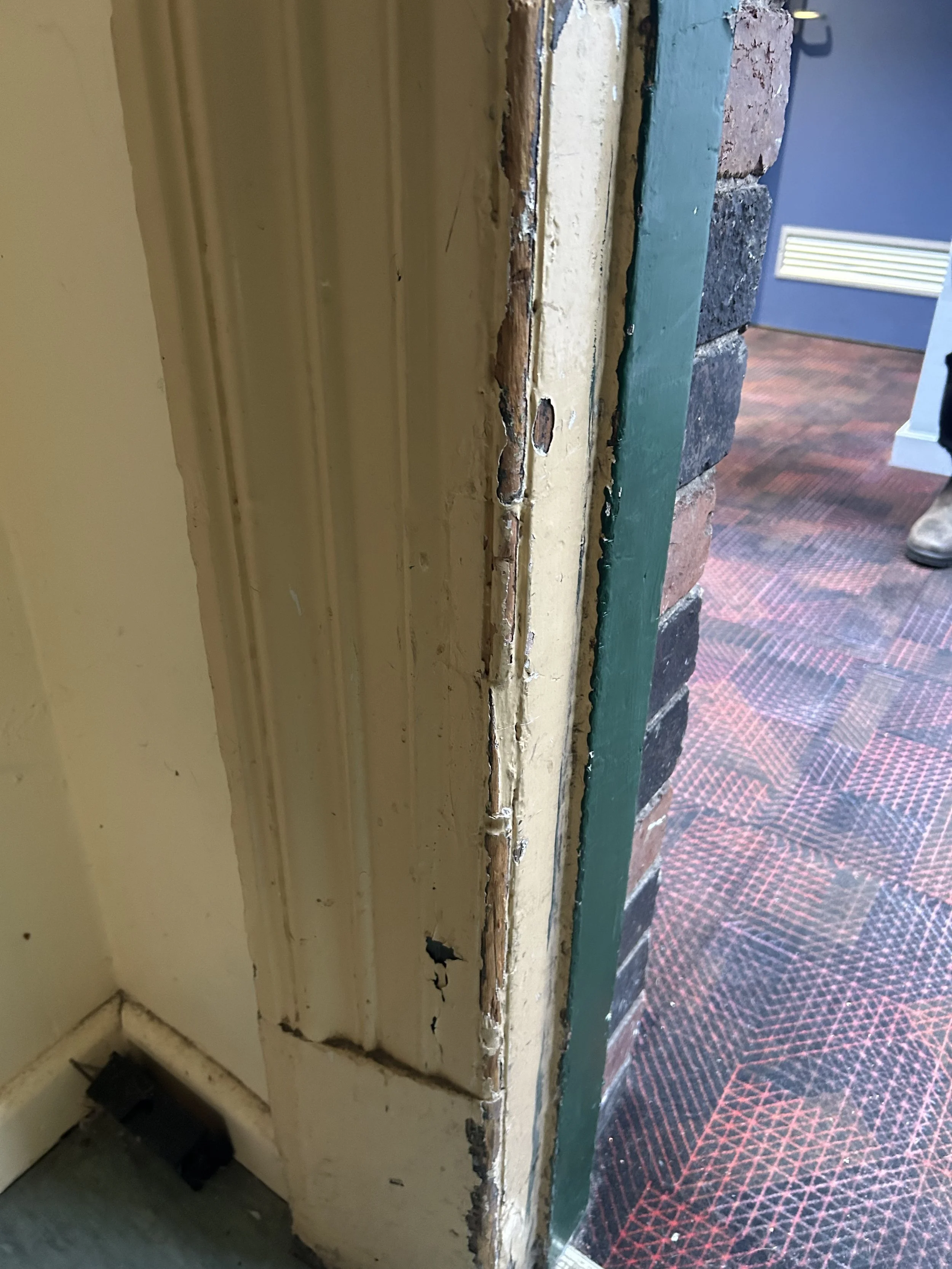 Close-up of a door frame with worn, chipped, and peeling paint revealing the wood underneath, next to a wall with a green trim and brick wall, with a carpeted floor.