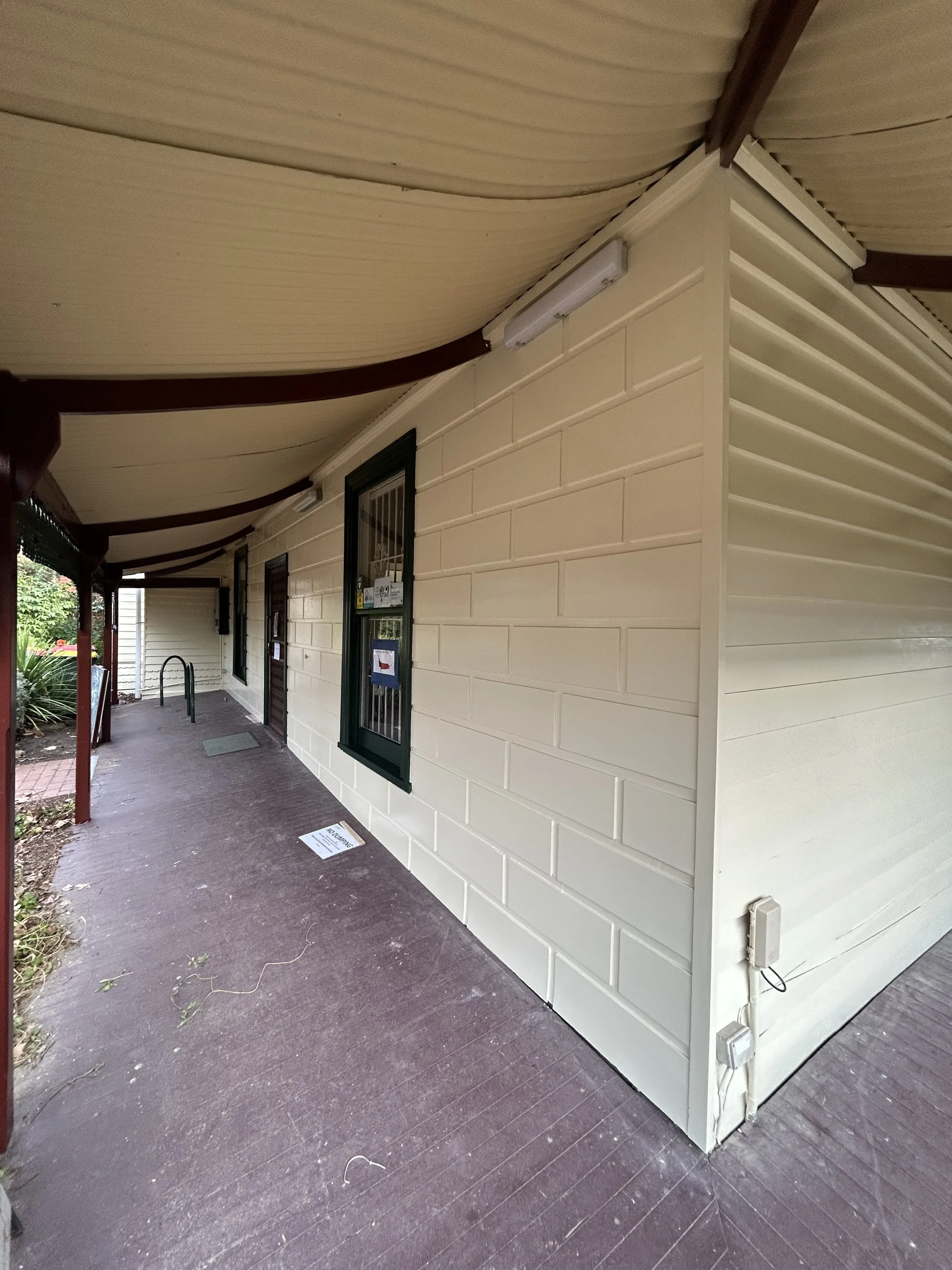 Residential building with cream-colored siding, green window frames, and a covered porch with a brown floor. Outdoor wiring and equipment are visible.