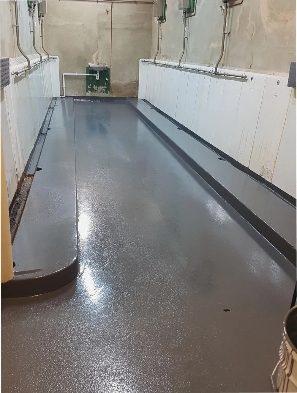 Empty industrial space with newly painted gray floor, pipes along the walls, and a small sink in the back corner.