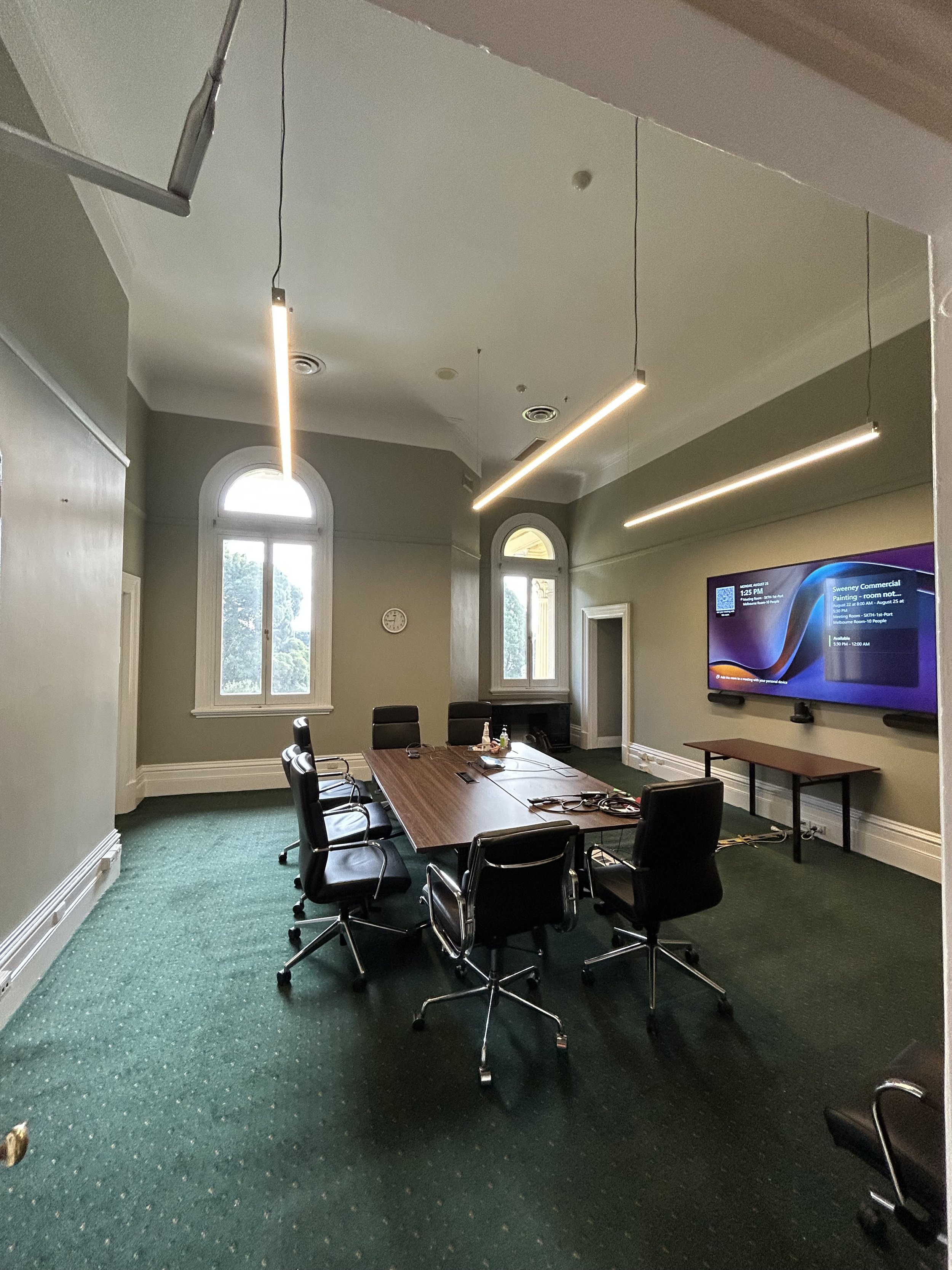Empty conference room with a large rectangular wooden table surrounded by black rolling chairs, green carpet, tall arched windows, a wall-mounted TV, and modern hanging lights.