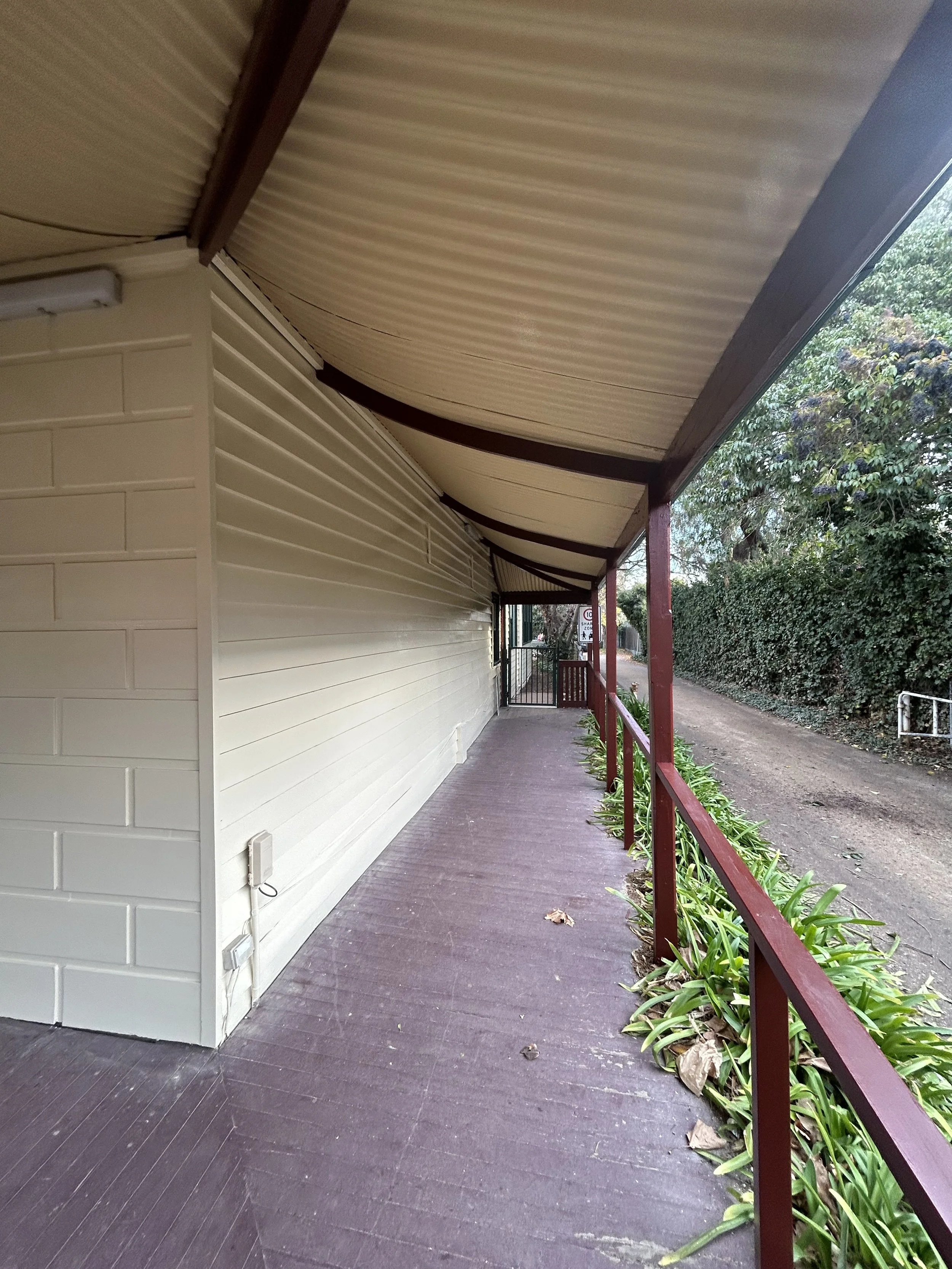 Empty porch with purple wooden flooring, beige plastic siding walls, a curved beige metal roof, red railing, green plants along the edge, and a dirt road to the right with bushes and trees.