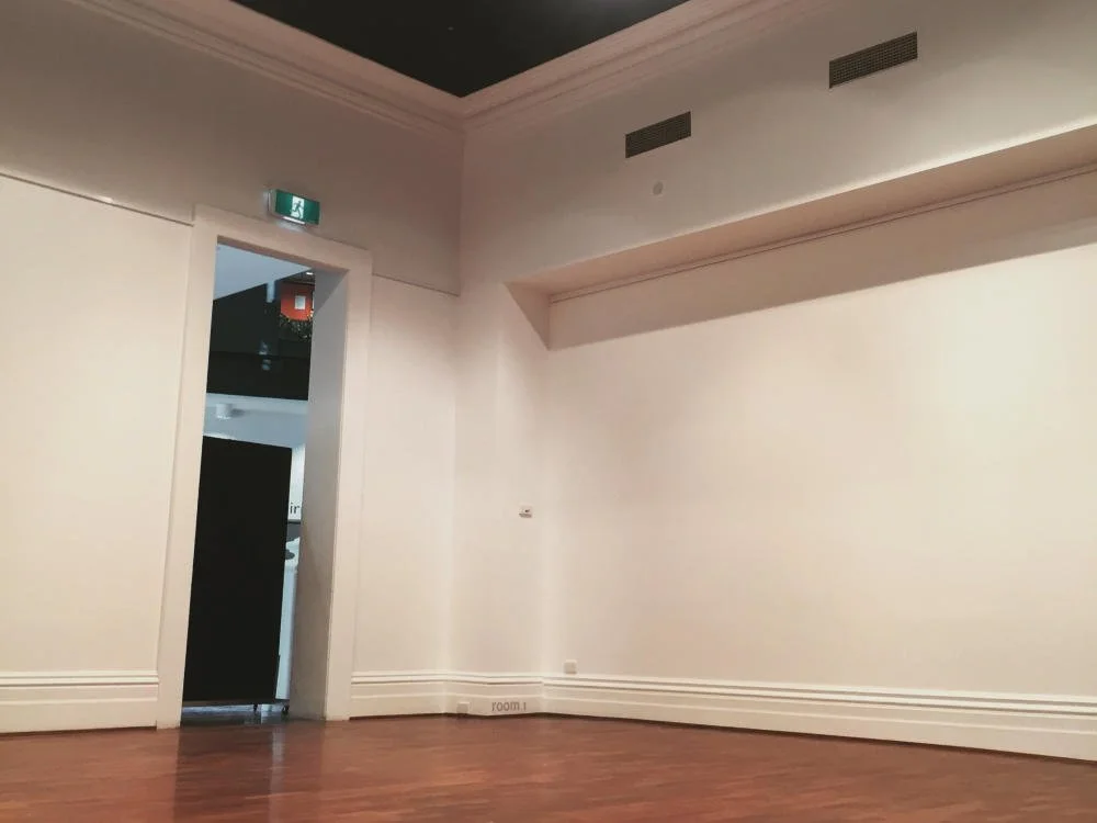 Empty room with white walls, hardwood floor, ceiling vents, and an open doorway leading to another room.