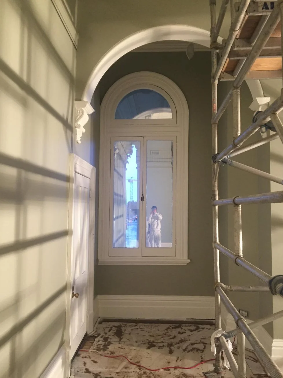 Interior room under renovation with a large arched window and a windowed door, revealing a person taking a photo outside, along with construction tools and scaffolding.