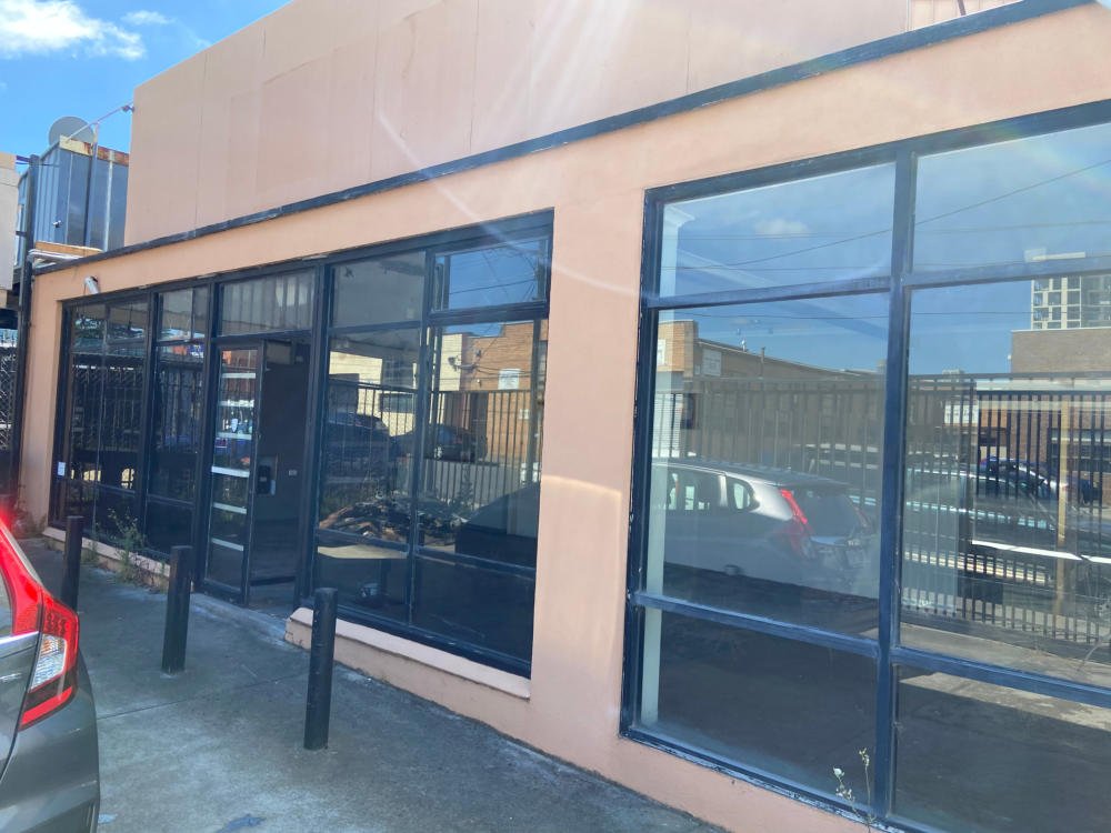 The exterior of a commercial building with large glass windows and a beige facade, parking lot with cars, and black bollards in front.