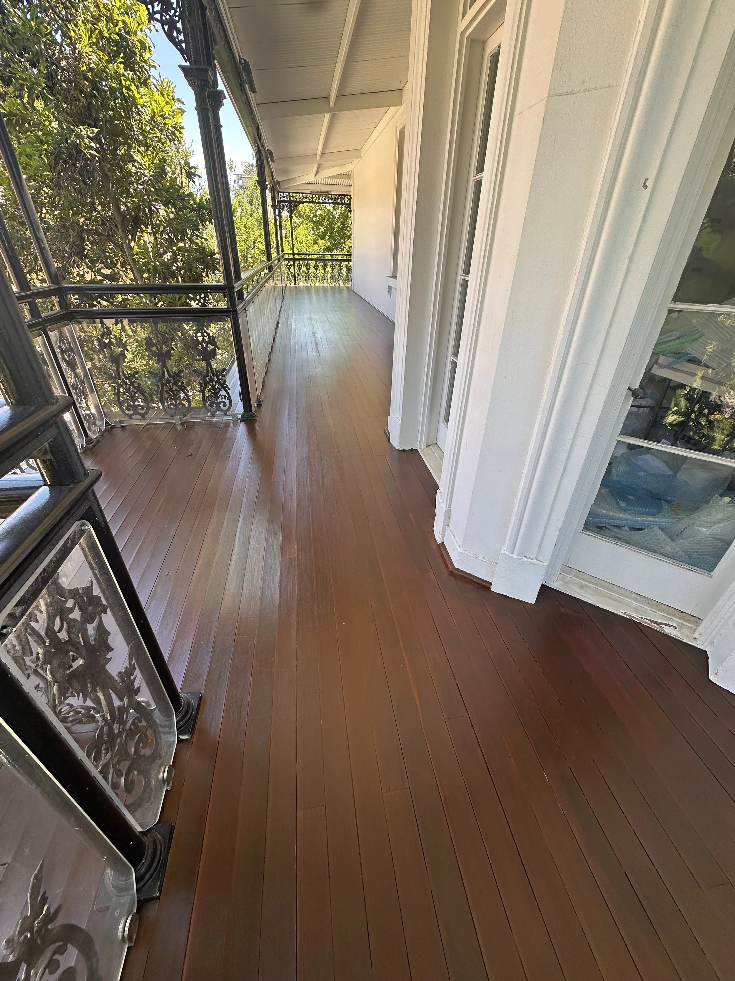 Porch with wooden flooring, black iron railings, white walls, and glass-paneled doors, surrounded by greenery.
