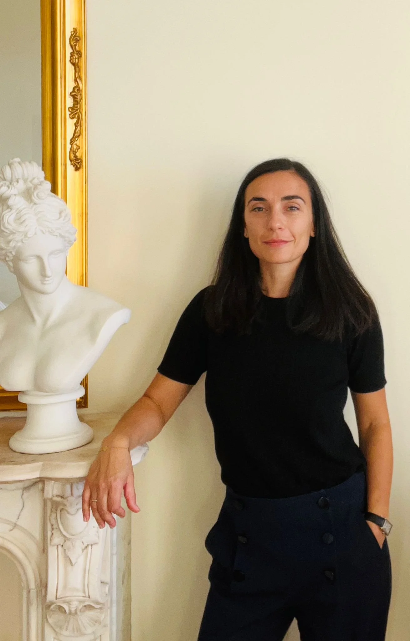 A woman with dark hair wearing a black shirt and navy pants standing next to a white bust sculpture on a marble fireplace with a gold-framed mirror behind her.