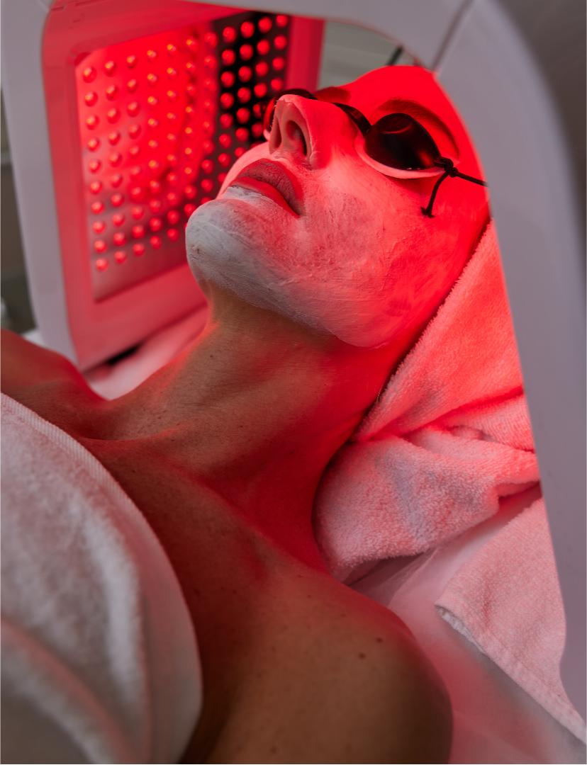 A person lying on a treatment bed wearing protective goggles, receiving red light therapy under a red light panel.