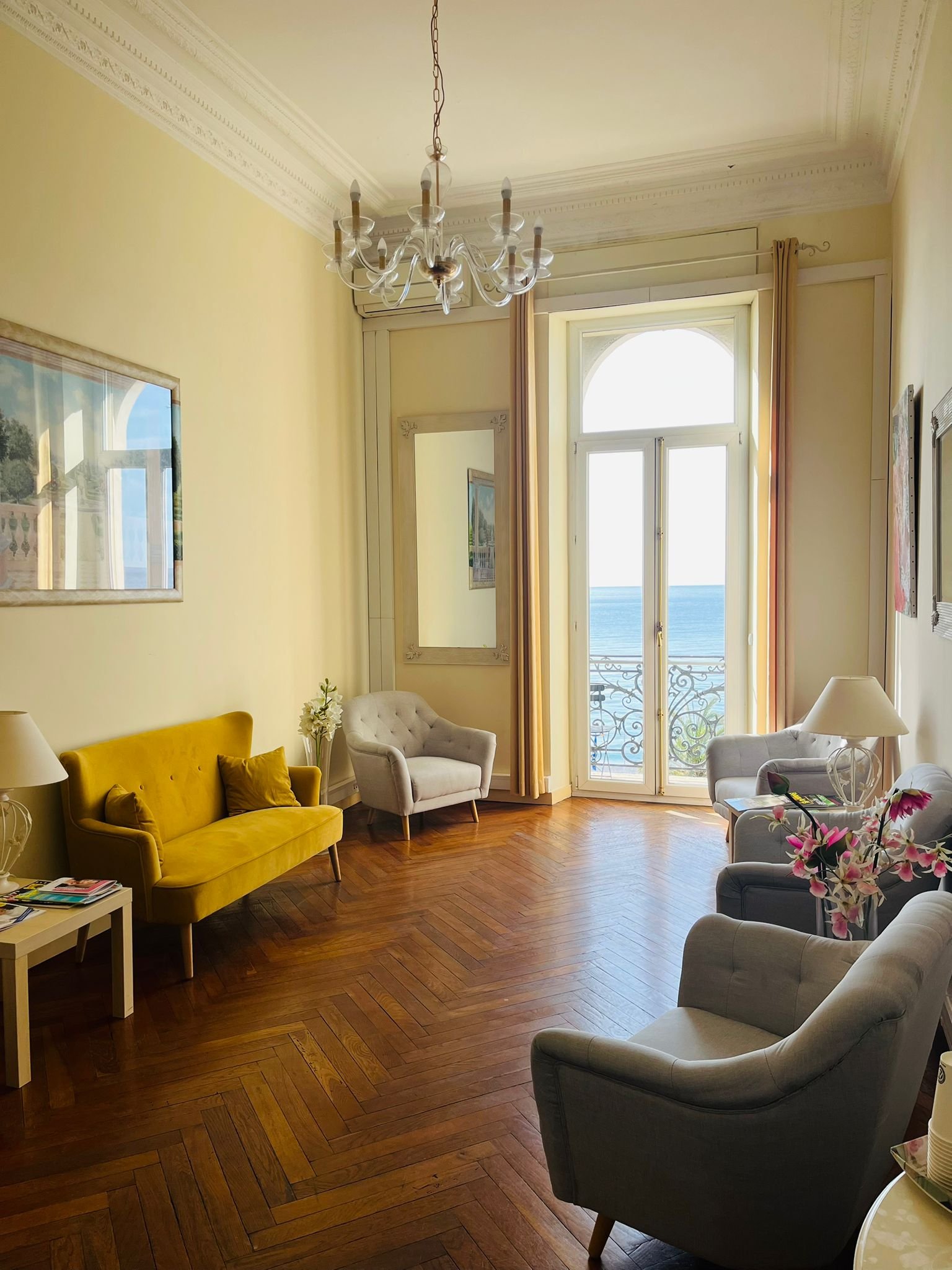 A living room with a balcony overlooking the ocean, featuring yellow and white sofas, armchairs, a chandelier, and decorative mirrors.