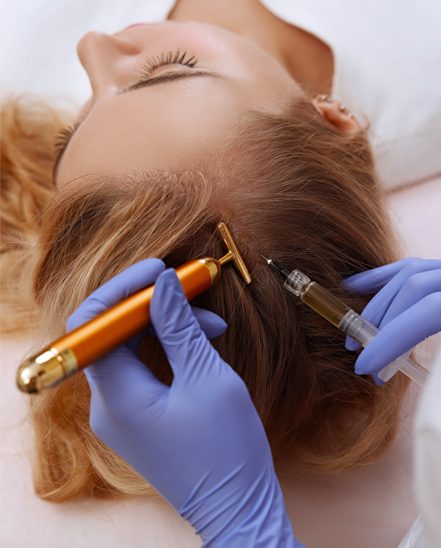 Close-up of a person receiving a hair injection procedure, lying down with their eyes closed. The technician, wearing blue gloves, holds a syringe near the person's scalp while another tool is being used on the scalp.