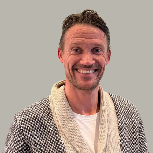 A man with short, wavy brown hair, smiling, wearing a patterned cardigan over a white shirt, standing against a plain light-colored wall.