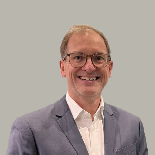 A smiling man wearing glasses, a white shirt, and a gray blazer against a plain background.