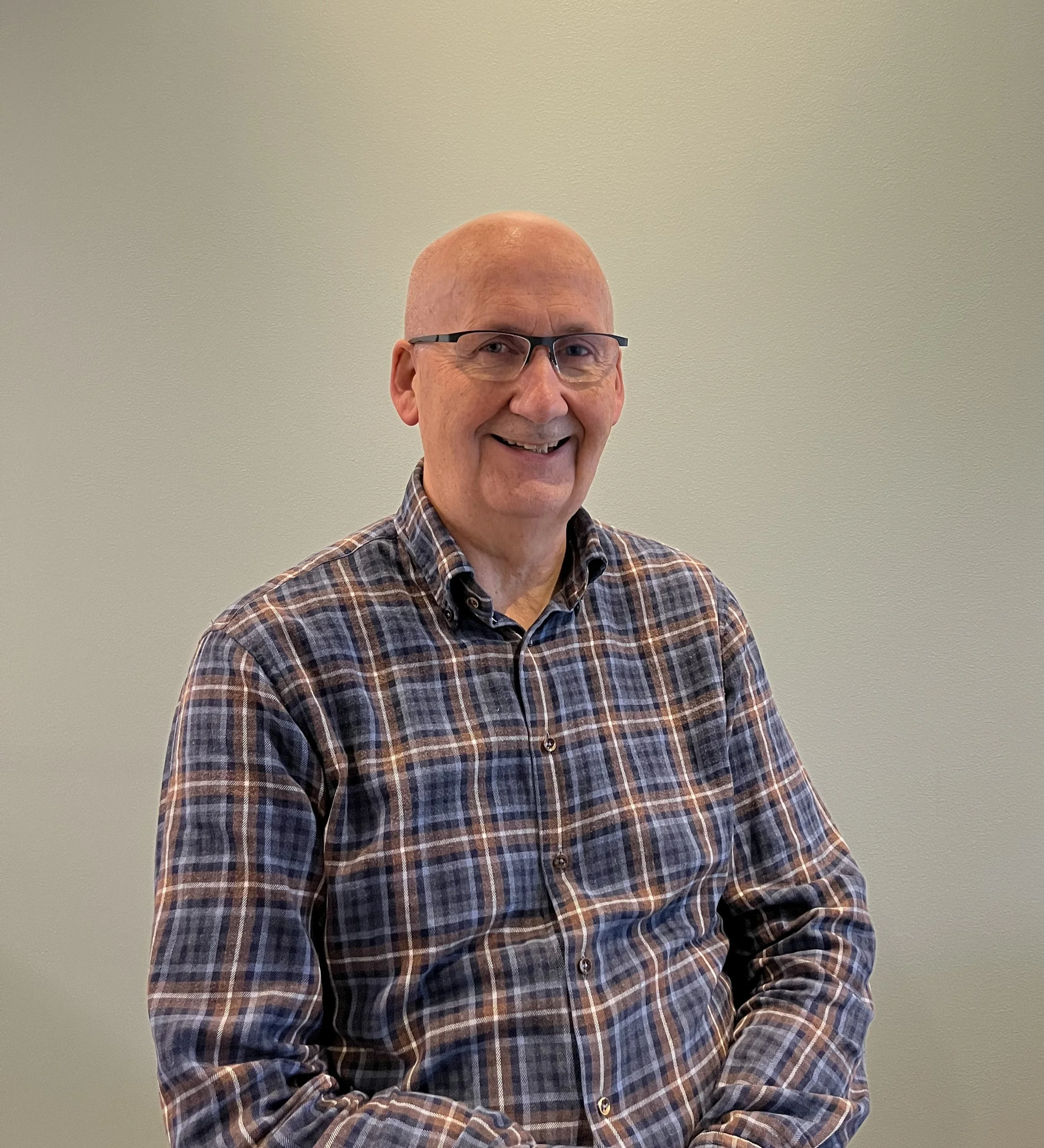 A smiling older man with glasses, wearing a plaid shirt, standing against a plain light-colored wall.