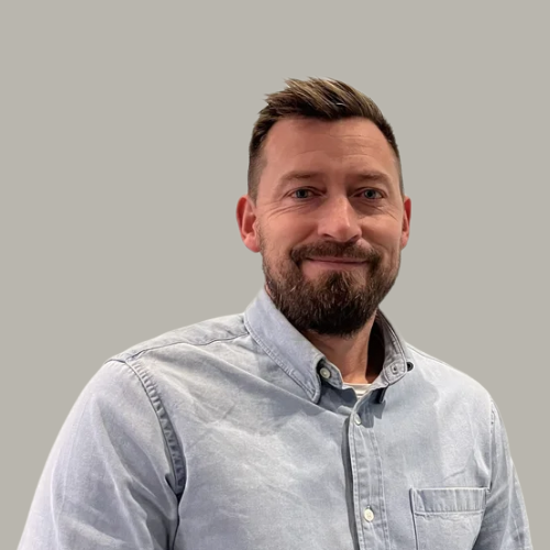 A man with a beard and short dark hair, wearing a light gray button-up shirt and a red watch, sitting against a plain light gray wall.