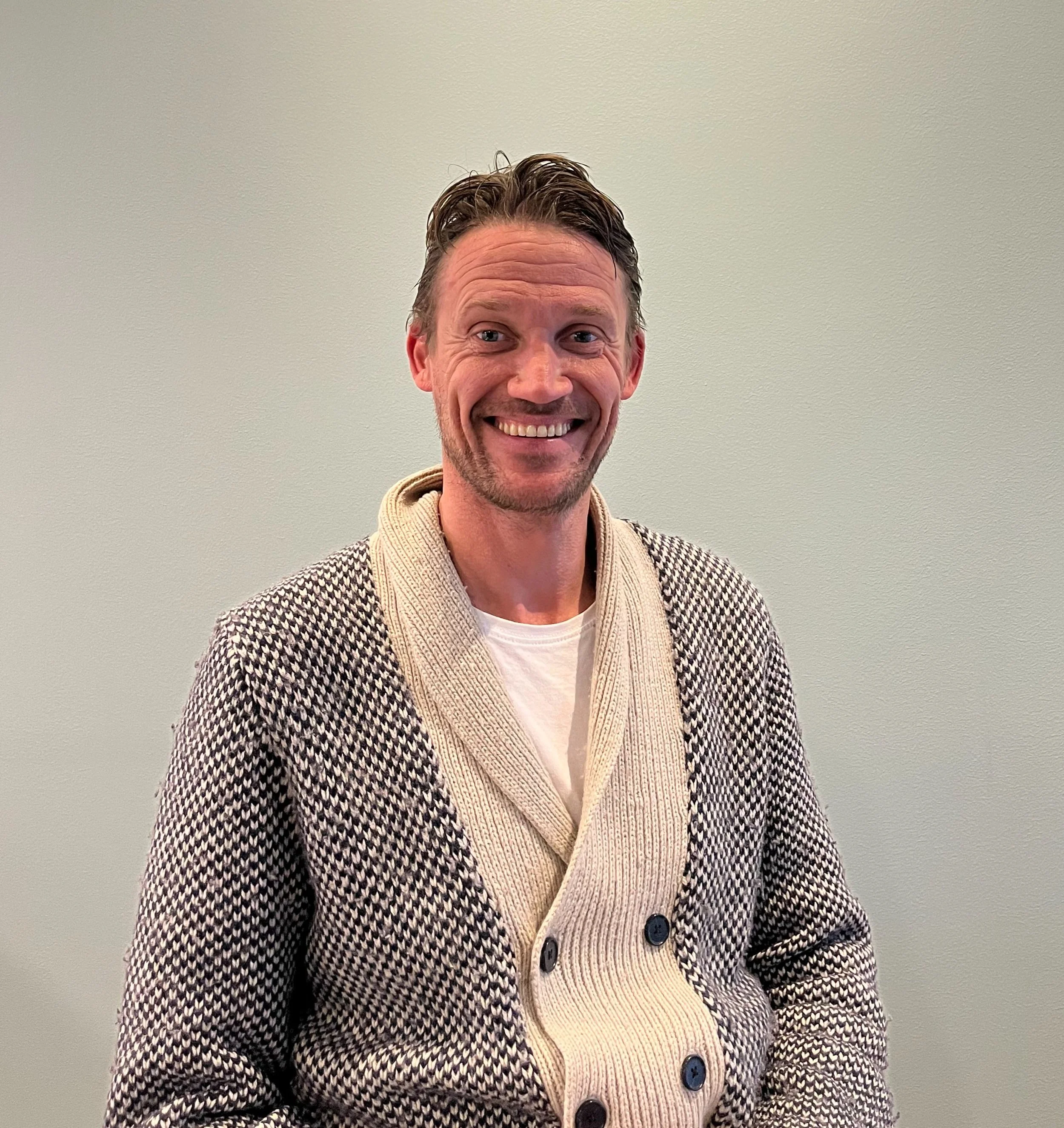 A man with short, wavy brown hair, smiling, wearing a patterned cardigan over a white shirt, standing against a plain light-colored wall.