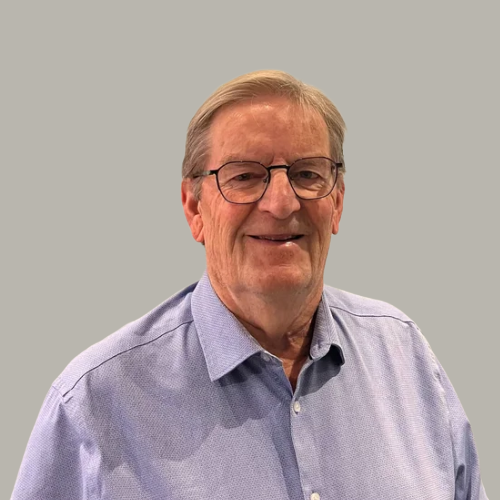 A middle-aged man with glasses and gray hair, wearing a light purple button-up shirt, standing in front of a plain light gray wall, smiling at the camera.