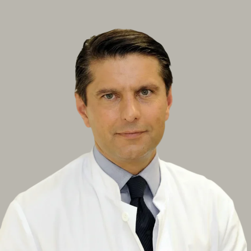 A man in a white medical coat and dark tie posing against a plain wall.