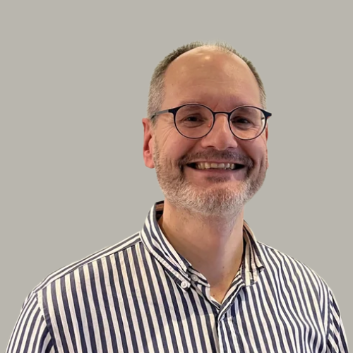 A smiling man with glasses and a beard, wearing a striped shirt, standing against a plain wall.