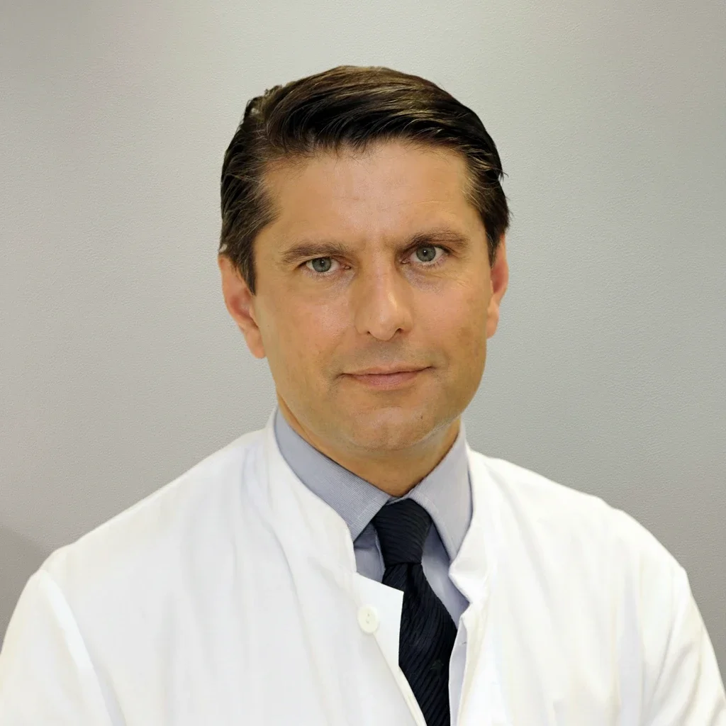 A man in a white medical coat and dark tie posing against a plain wall.