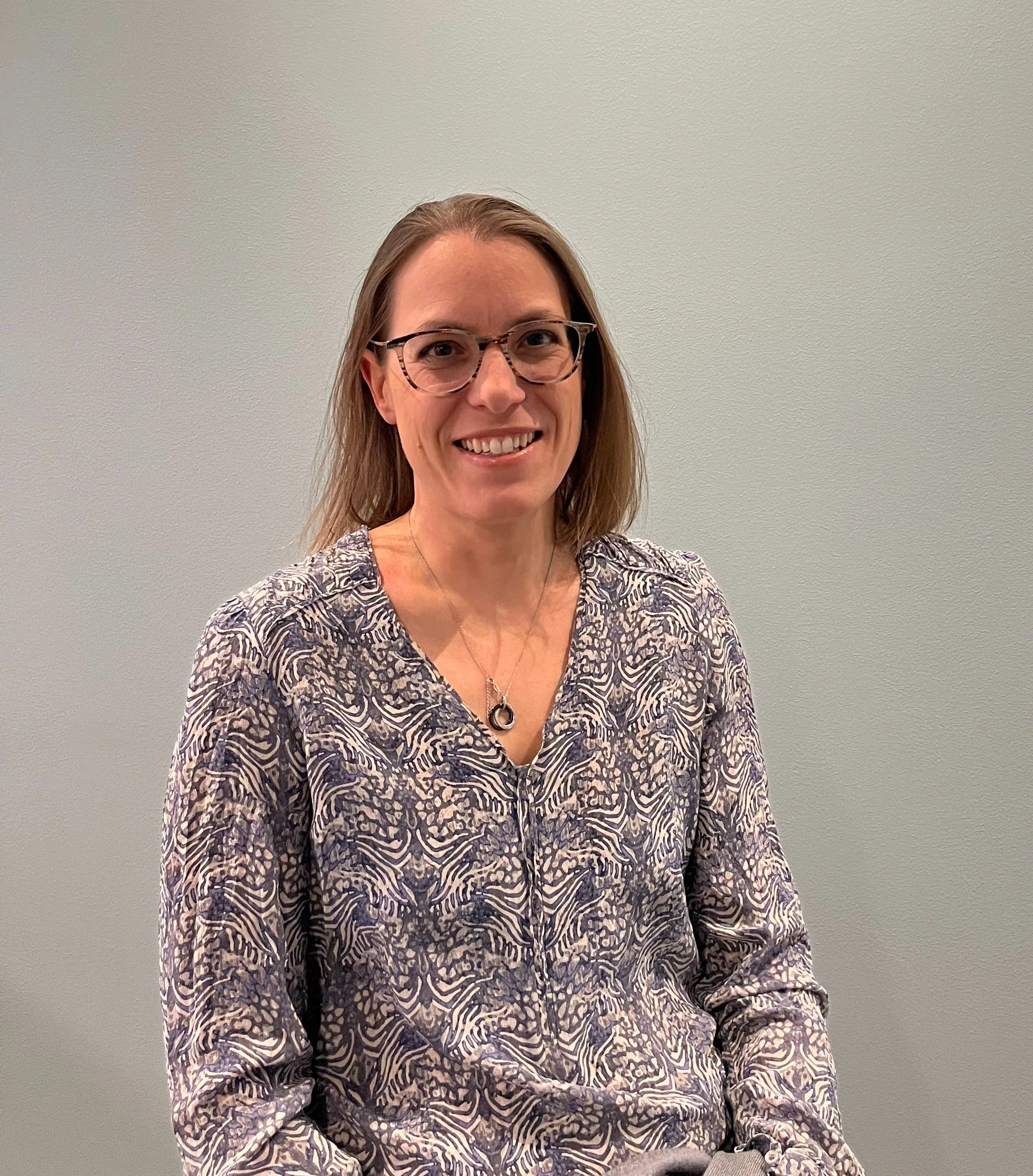 A woman with glasses smiles at the camera against a plain wall.