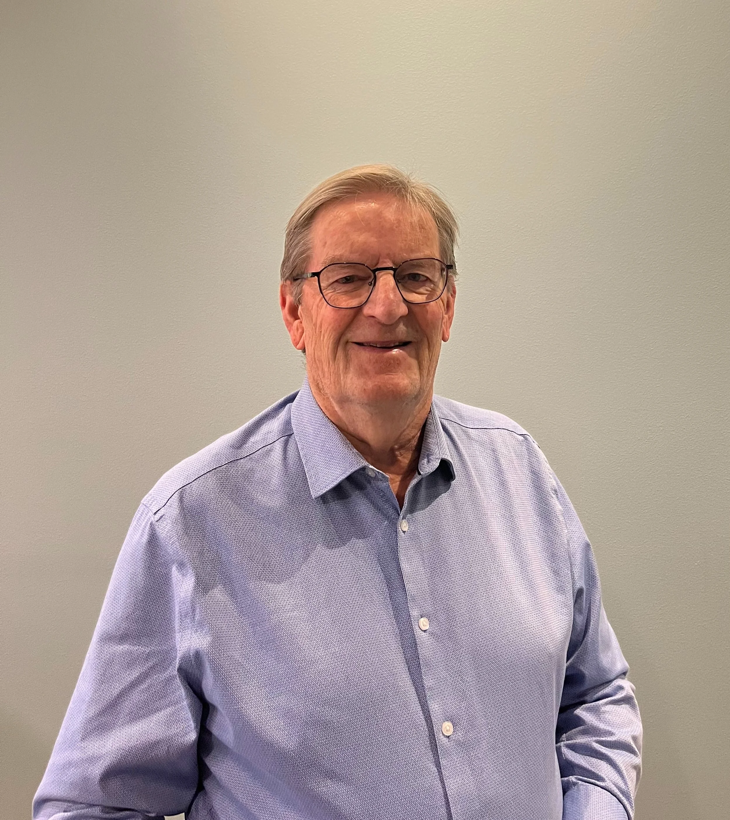 A middle-aged man with glasses and gray hair, wearing a light purple button-up shirt, standing in front of a plain light gray wall, smiling at the camera.