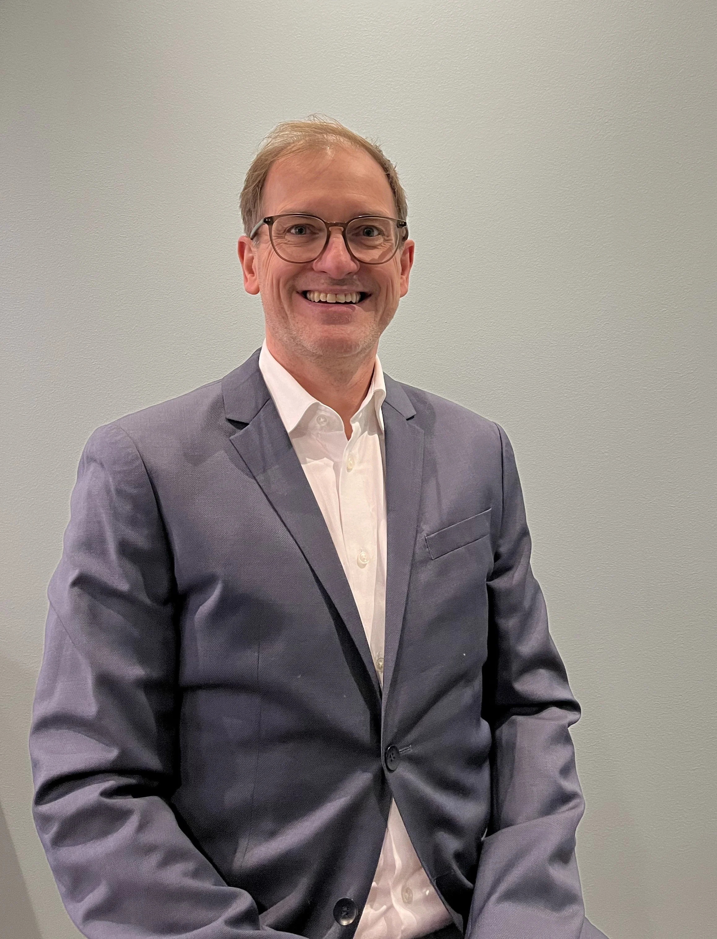 A smiling man wearing glasses, a white shirt, and a gray blazer against a plain background.