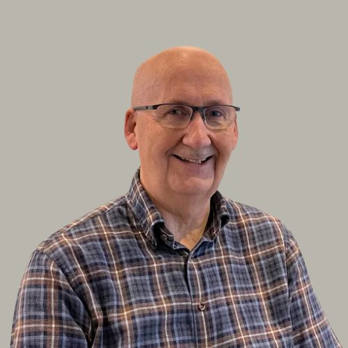A smiling older man with glasses, wearing a plaid shirt, standing against a plain light-colored wall.