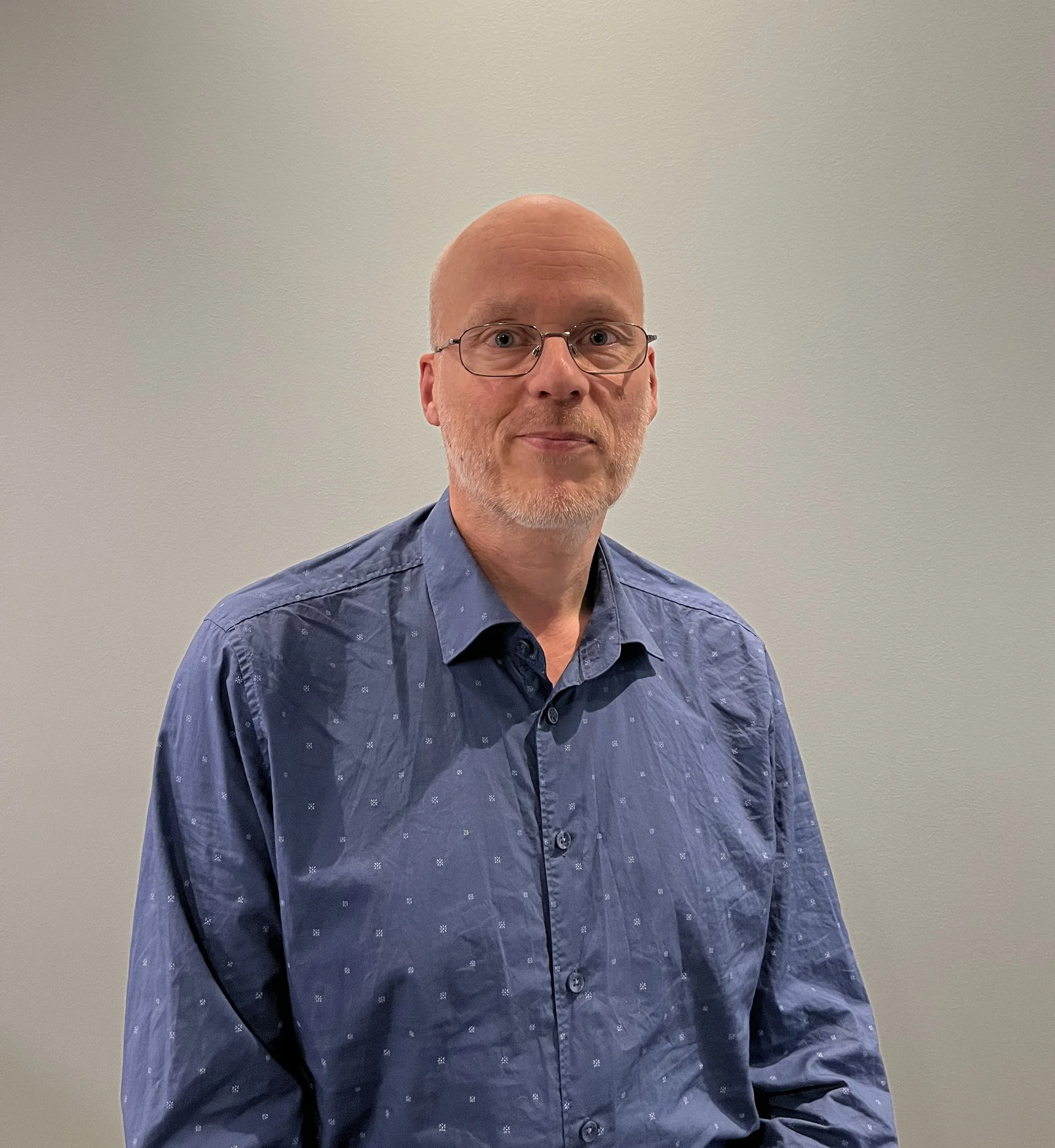 A middle-aged man with glasses, a bald head, and facial hair, wearing a blue patterned button-up shirt, standing against a plain gray wall.
