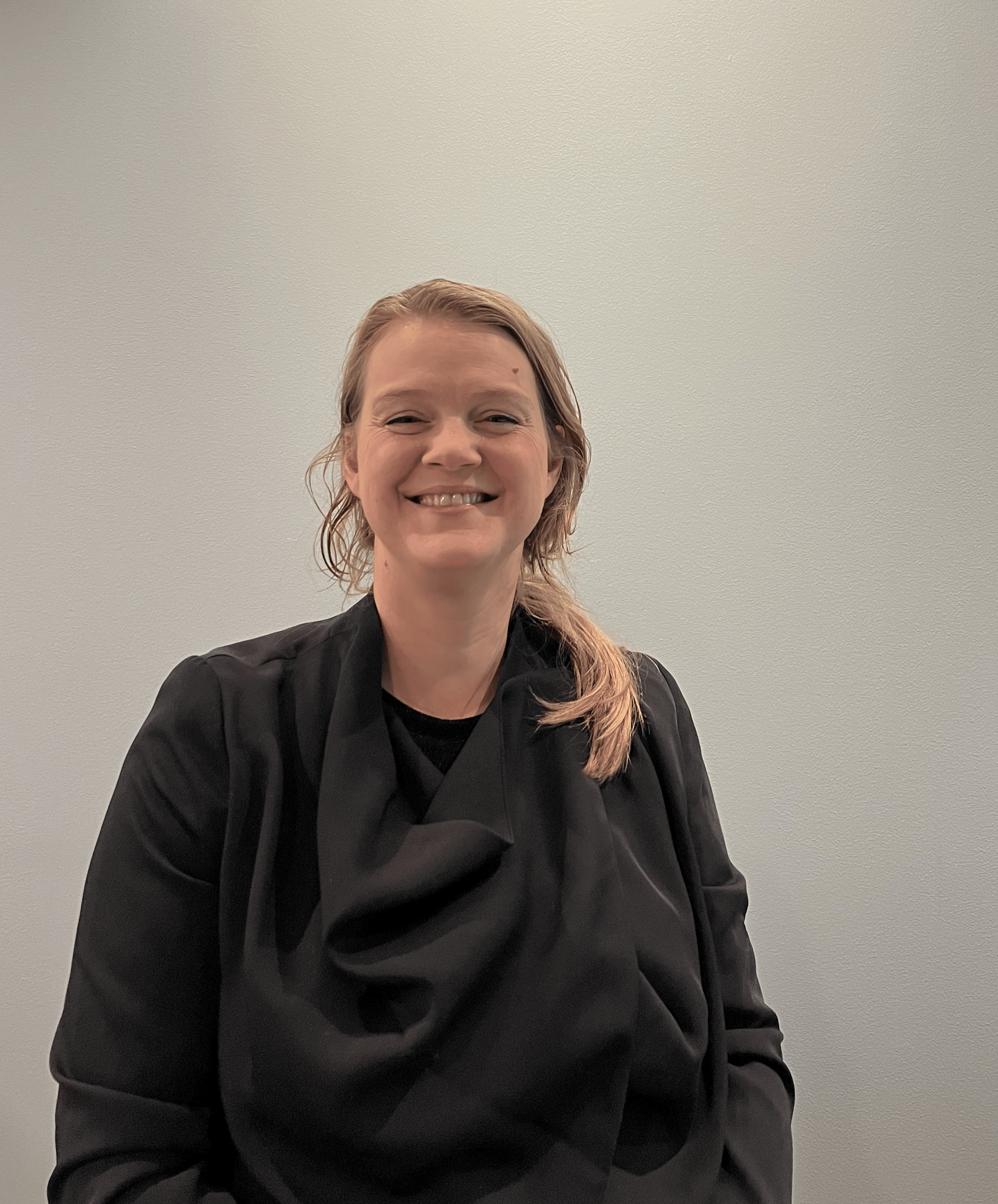 Smiling woman with light brown, wavy hair wearing a black blazer standing against a plain, off-white wall.