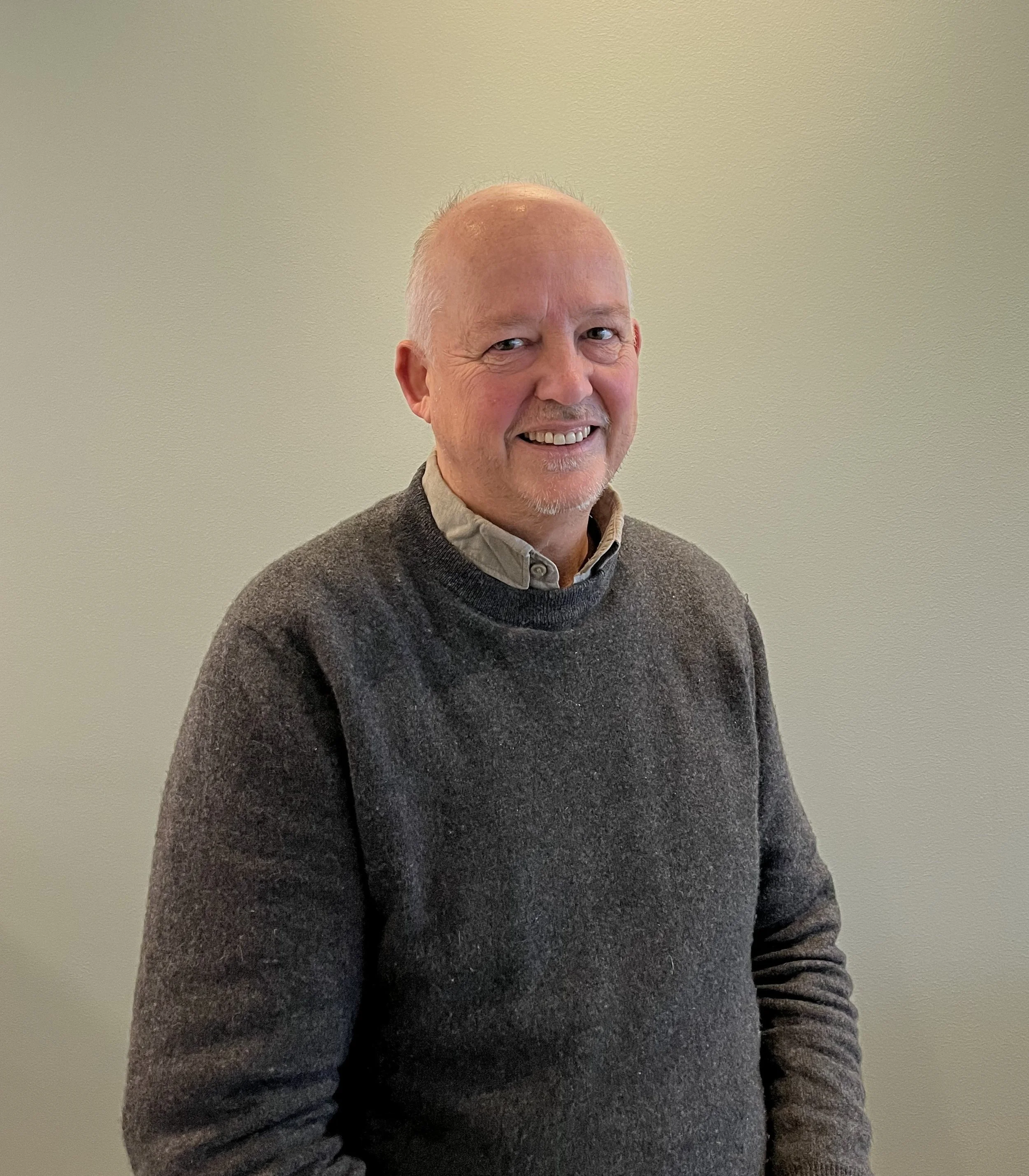 An older man with light skin, short gray hair, and a goatee smiling at the camera, wearing a dark gray sweater over a light collared shirt, standing against a plain light-colored background.