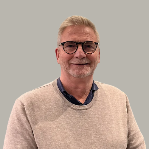 A middle-aged man with light hair, glasses, and a beard, smiling at the camera, wearing a beige sweater over a button-up shirt, standing against a plain, light-colored wall.