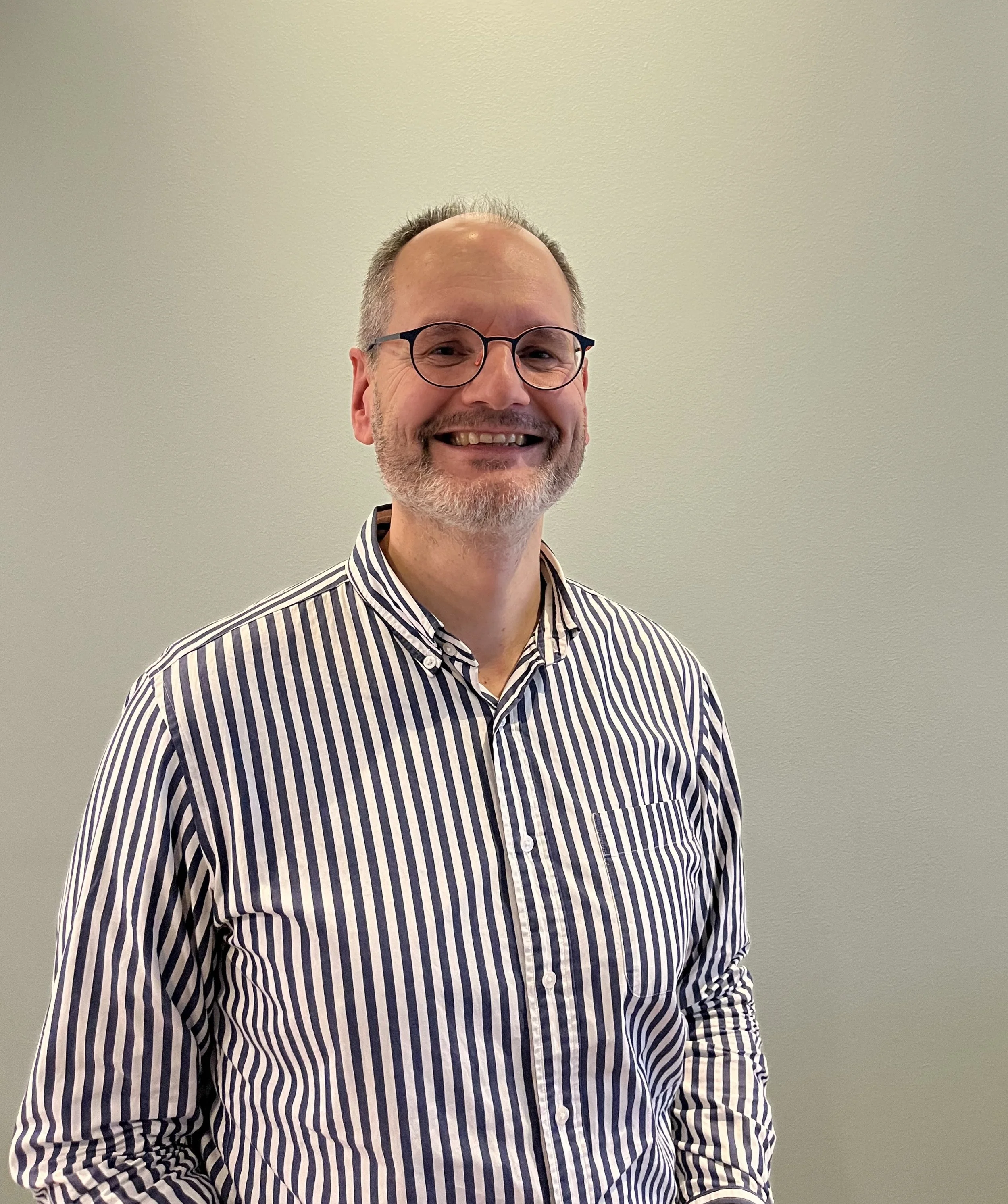 A smiling man with glasses and a beard, wearing a striped shirt, standing against a plain wall.
