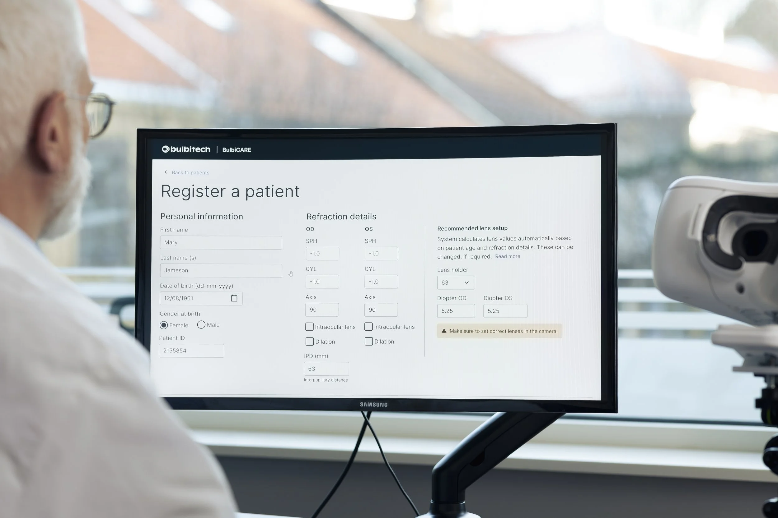 A medical professional looking at a computer monitor displaying a patient registration form with personal and refraction details in a clinical setting with medical equipment nearby.
