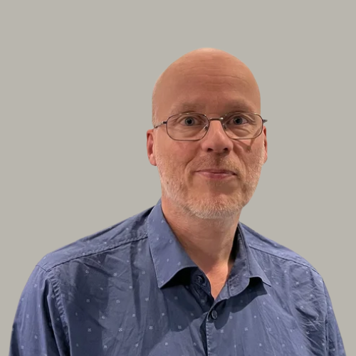 A middle-aged man with glasses, a bald head, and facial hair, wearing a blue patterned button-up shirt, standing against a plain gray wall.