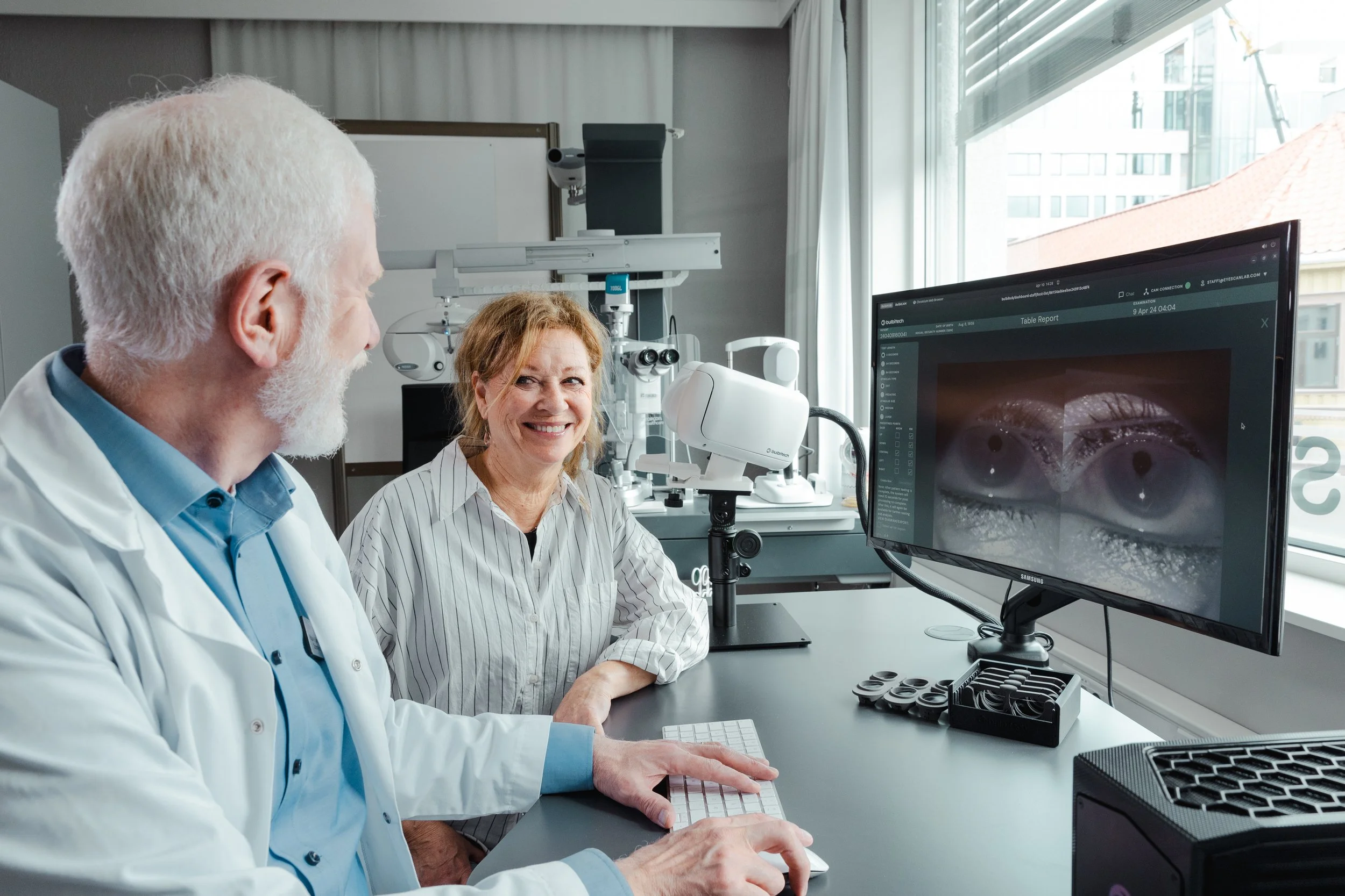 Two eye doctors in a modern clinic, with one examining a patient and the other monitoring images of eyes on a large computer screen.